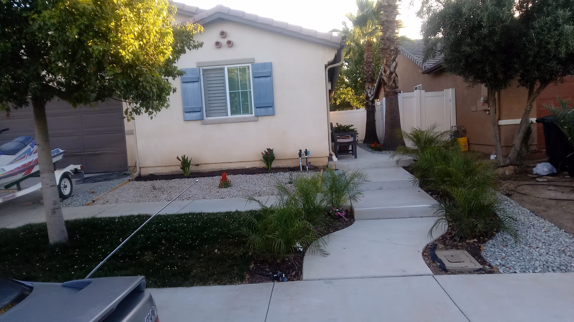 Exterior view of a single-story building with beige walls and blue window shutters. A concrete pathway leads up to the entrance, flanked by small palm plants and landscaping with gravel and grass. There is a tree on the left side and a boat parked near a garage door. Another building is partially visible on the right side.