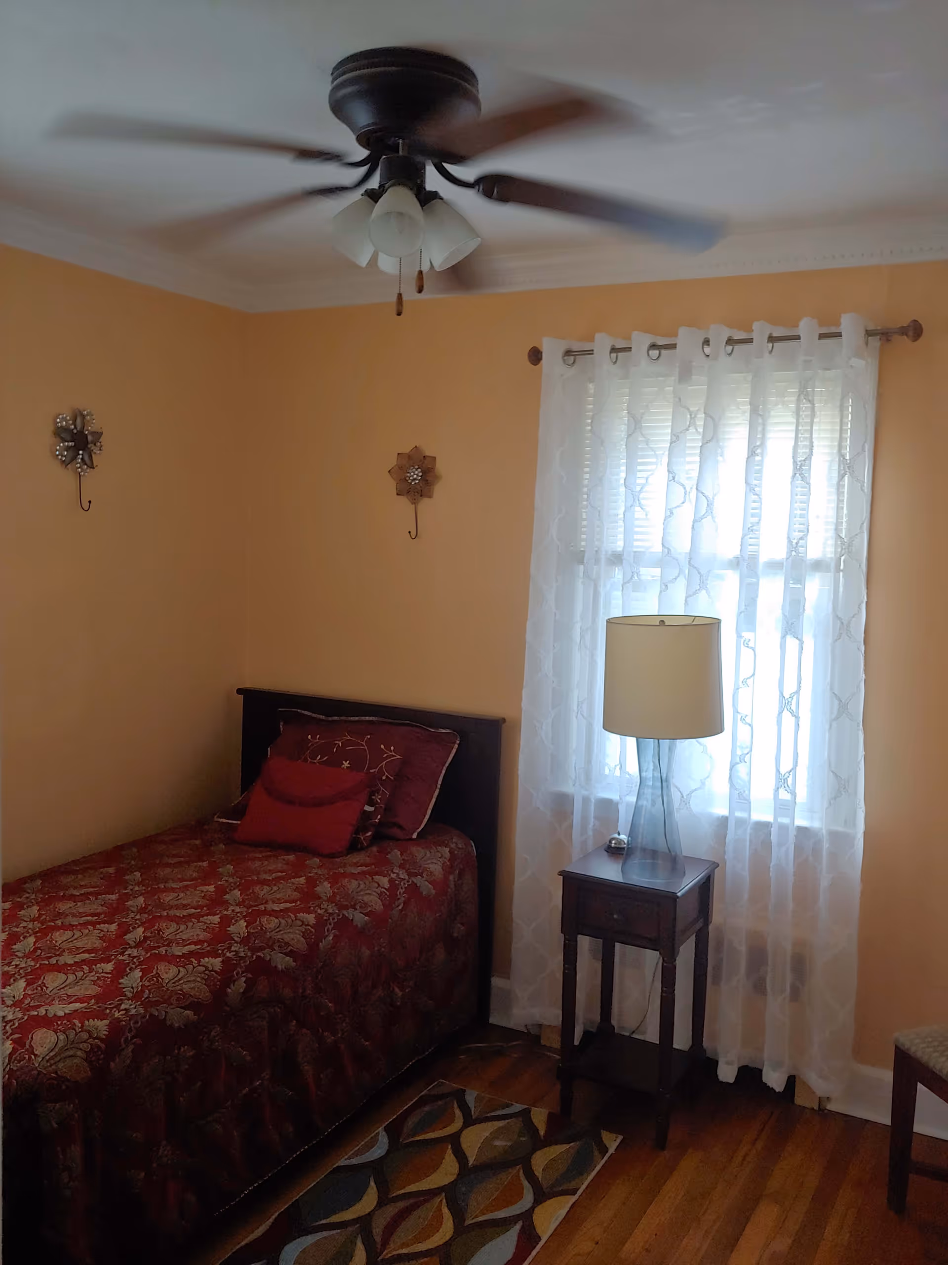 Cozy bedroom with a single bed covered in red patterned bedding, a nightstand with a lamp next to a window with sheer curtains, and a ceiling fan overhead.