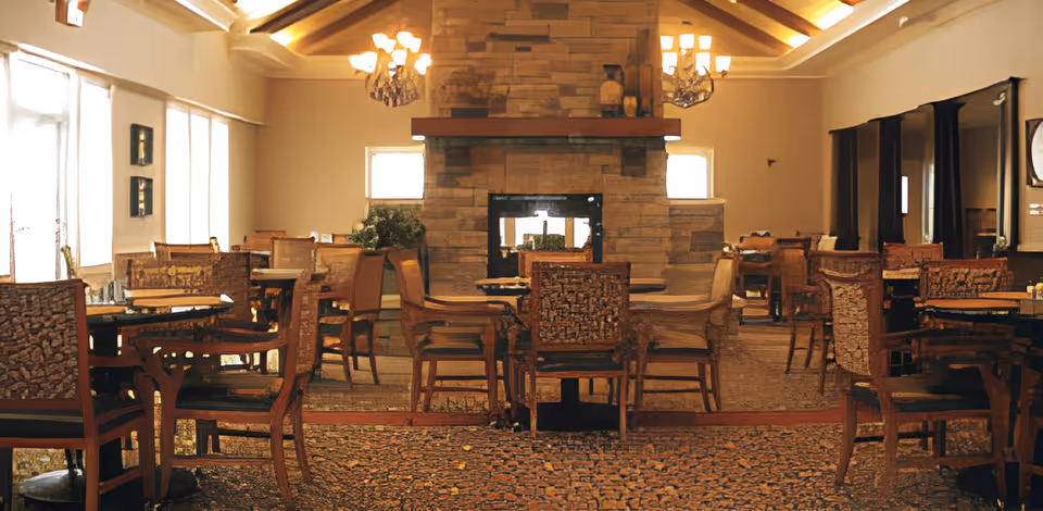 A warm and inviting dining room with multiple wooden tables and chairs arranged around a central stone fireplace. The room features large windows on one side allowing natural light to fill the space, chandeliers hanging from a vaulted ceiling, and a patterned carpet covering the floor.