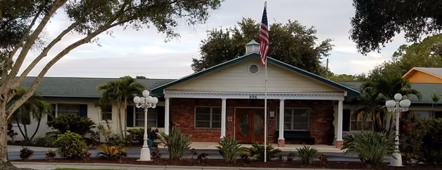 Front facade of a single-story brick building with a covered entrance, American flag on a pole, columns and landscaped shrubs.