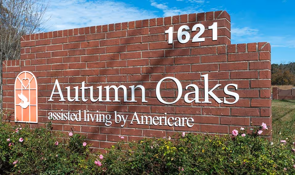 A brick sign for Autumn Oaks assisted living by Americare with the address number 1621 displayed on top. The sign is surrounded by green bushes with pink flowers and set against a blue sky background.