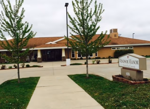 Front entrance of Windsor Manor senior living building with a driveway, trees, and a sign reading 'Windsor Manor'.