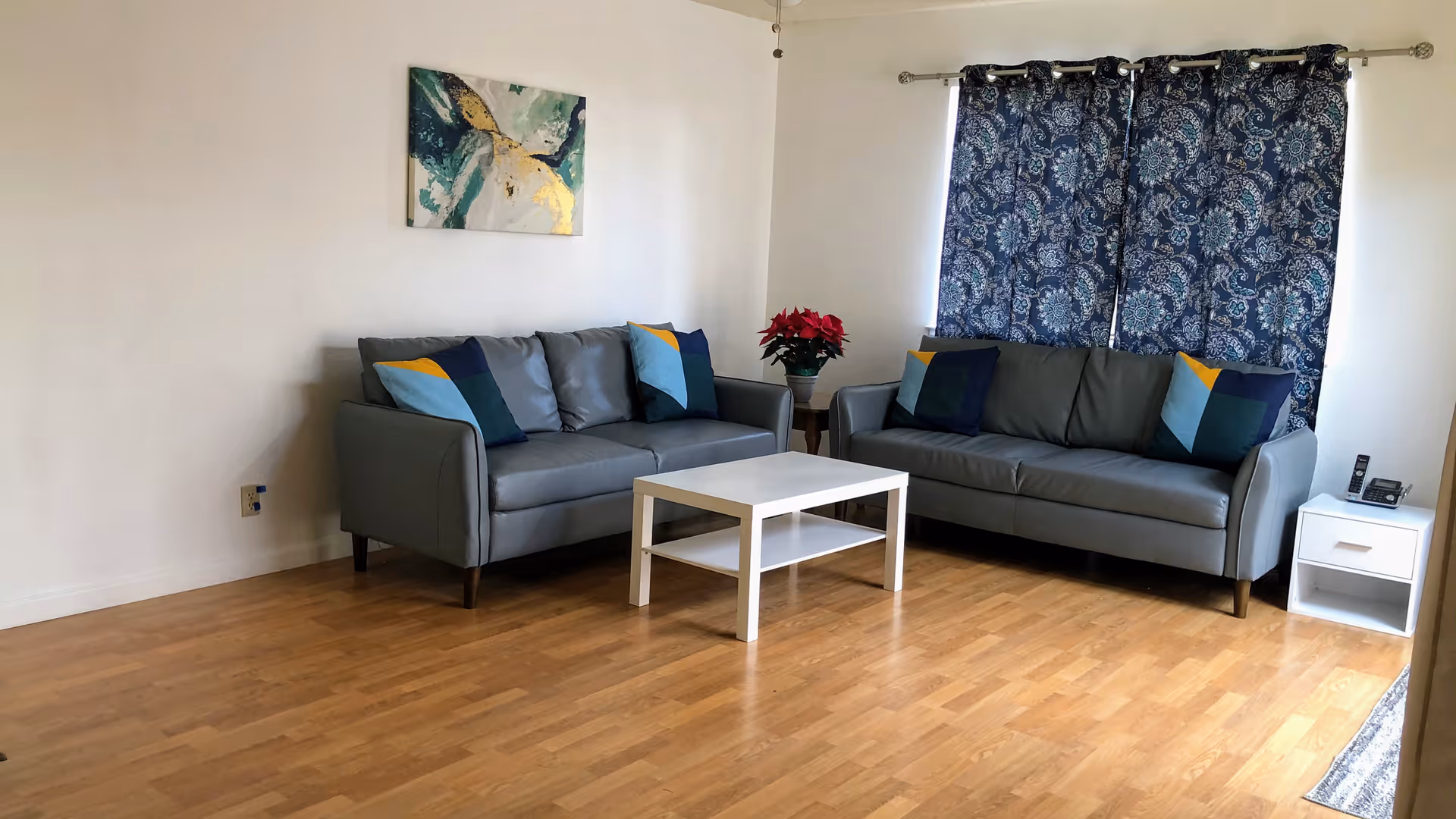 A living room with two gray sofas adorned with colorful throw pillows, a white coffee table in the center, a small side table with a red poinsettia plant, blue patterned curtains covering a window, and a modern abstract painting on the wall.