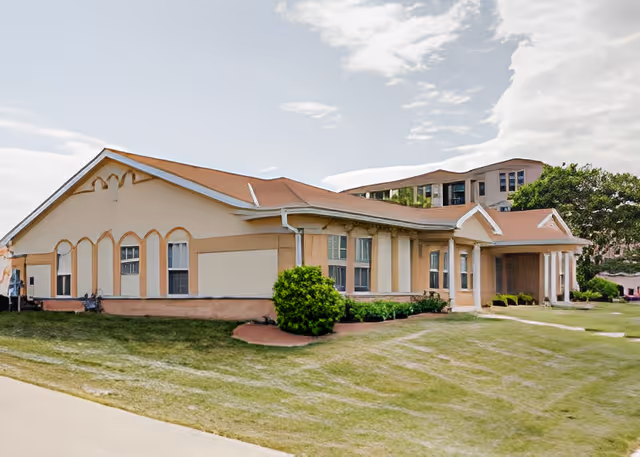 Front exterior of a single-story beige assisted living building with a covered columned entrance and surrounding lawn.