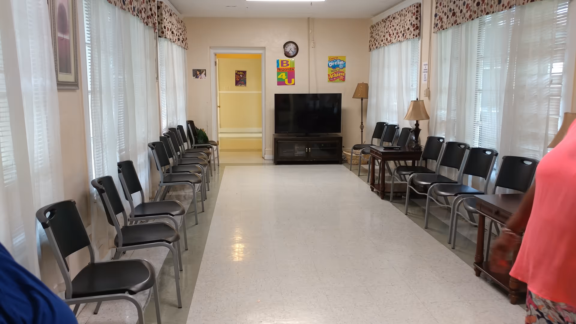 Hall-style living room in a senior facility with chairs along the walls and a TV.