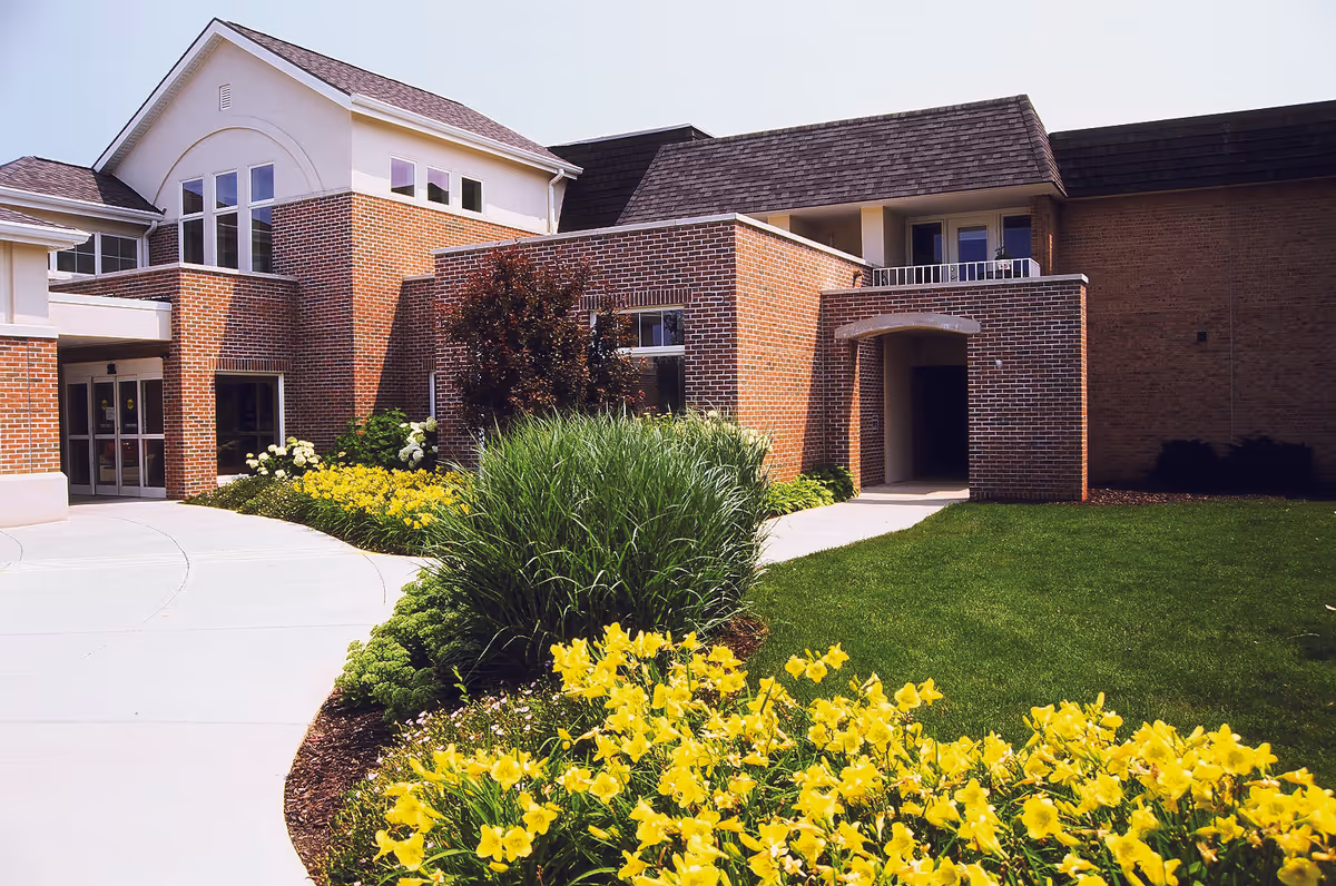 Brick exterior of a senior living facility with landscaped walkway, shrubs, and yellow flowers.