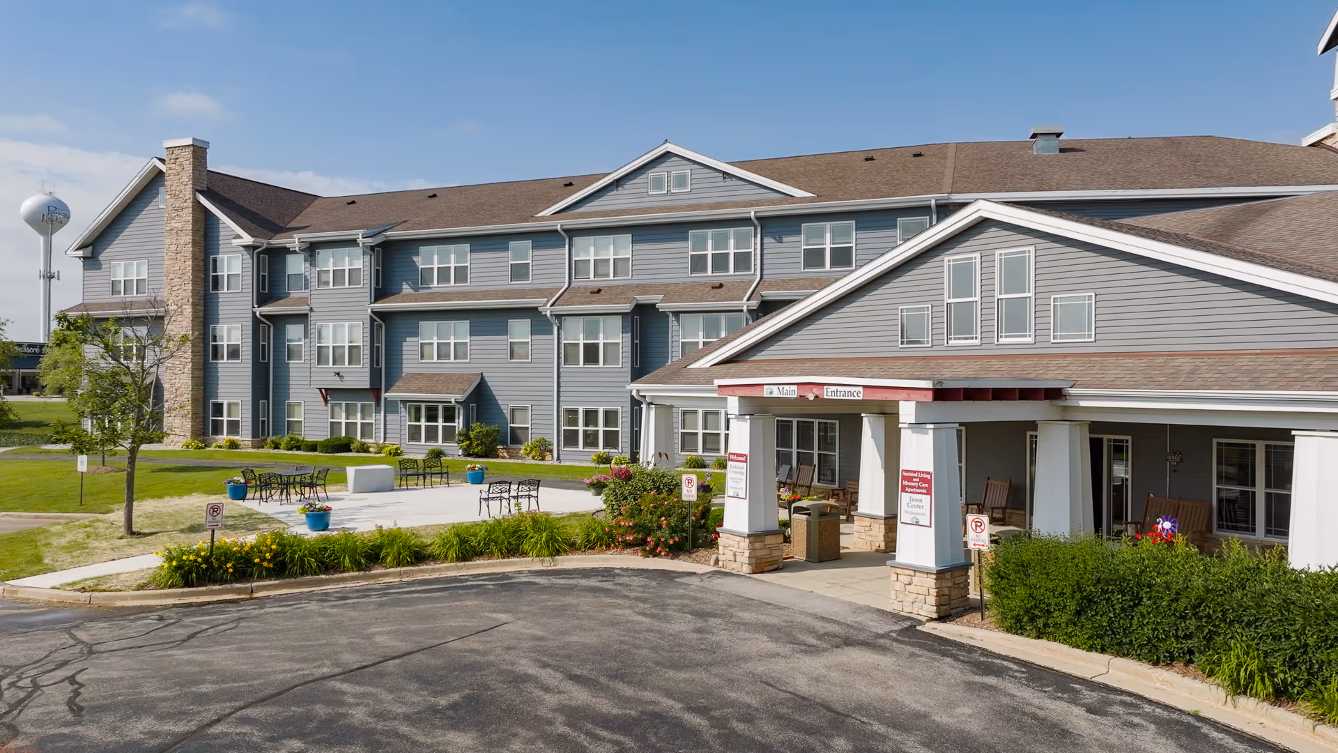 Exterior view of Kirkland Crossings senior living facility showing a large three-story building with gray siding and multiple windows. The entrance has a covered porch with white pillars and stone bases, labeled 'Main Entrance'. There is a paved driveway and a small patio area with tables, chairs, and potted plants. A water tower is visible in the background under a partly cloudy sky.