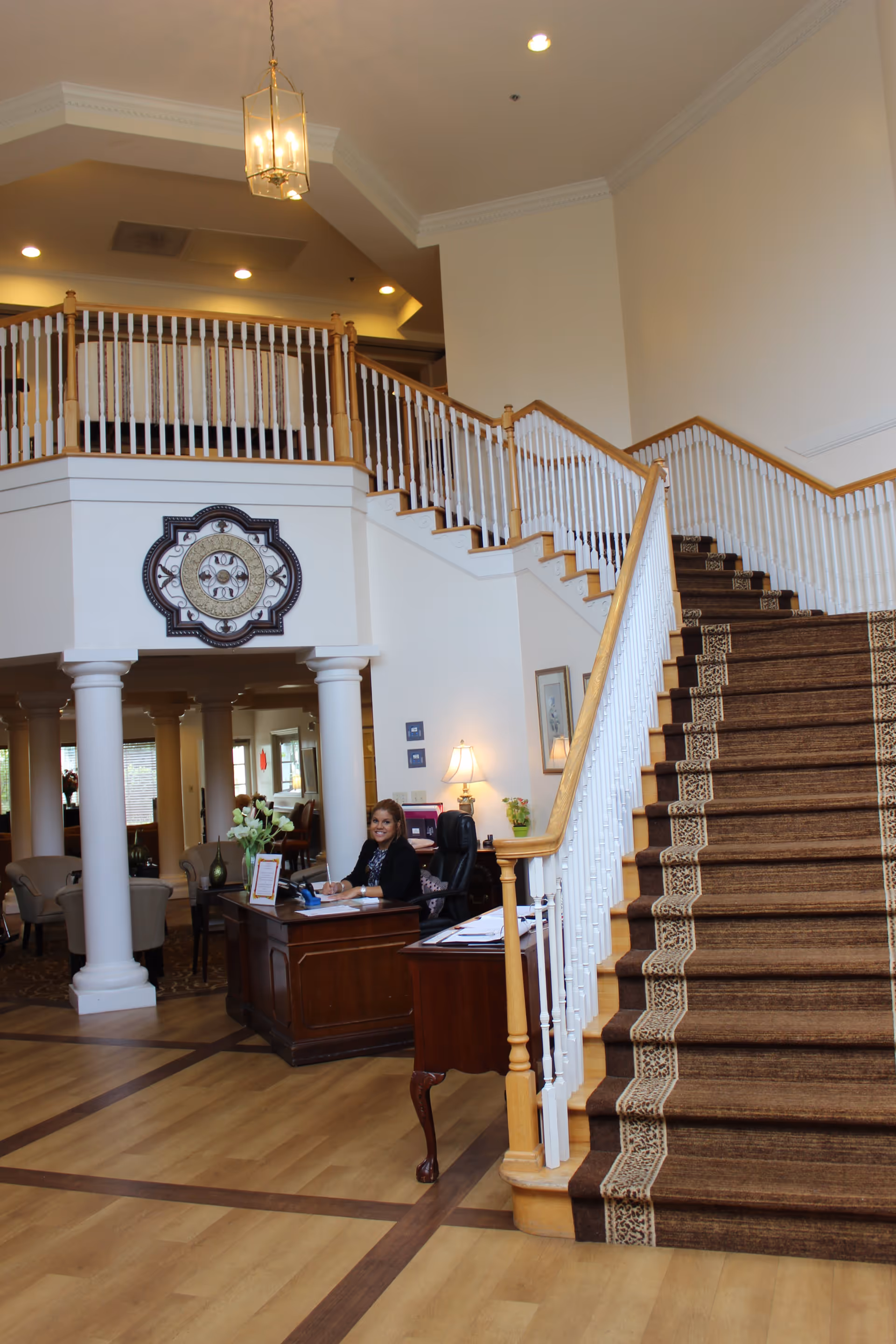 Interior view of a senior living facility lobby with a large staircase covered in a brown patterned carpet. A woman is seated at a wooden reception desk with flowers and paperwork. The area features white columns, wooden flooring, and a decorative wall clock above the desk.