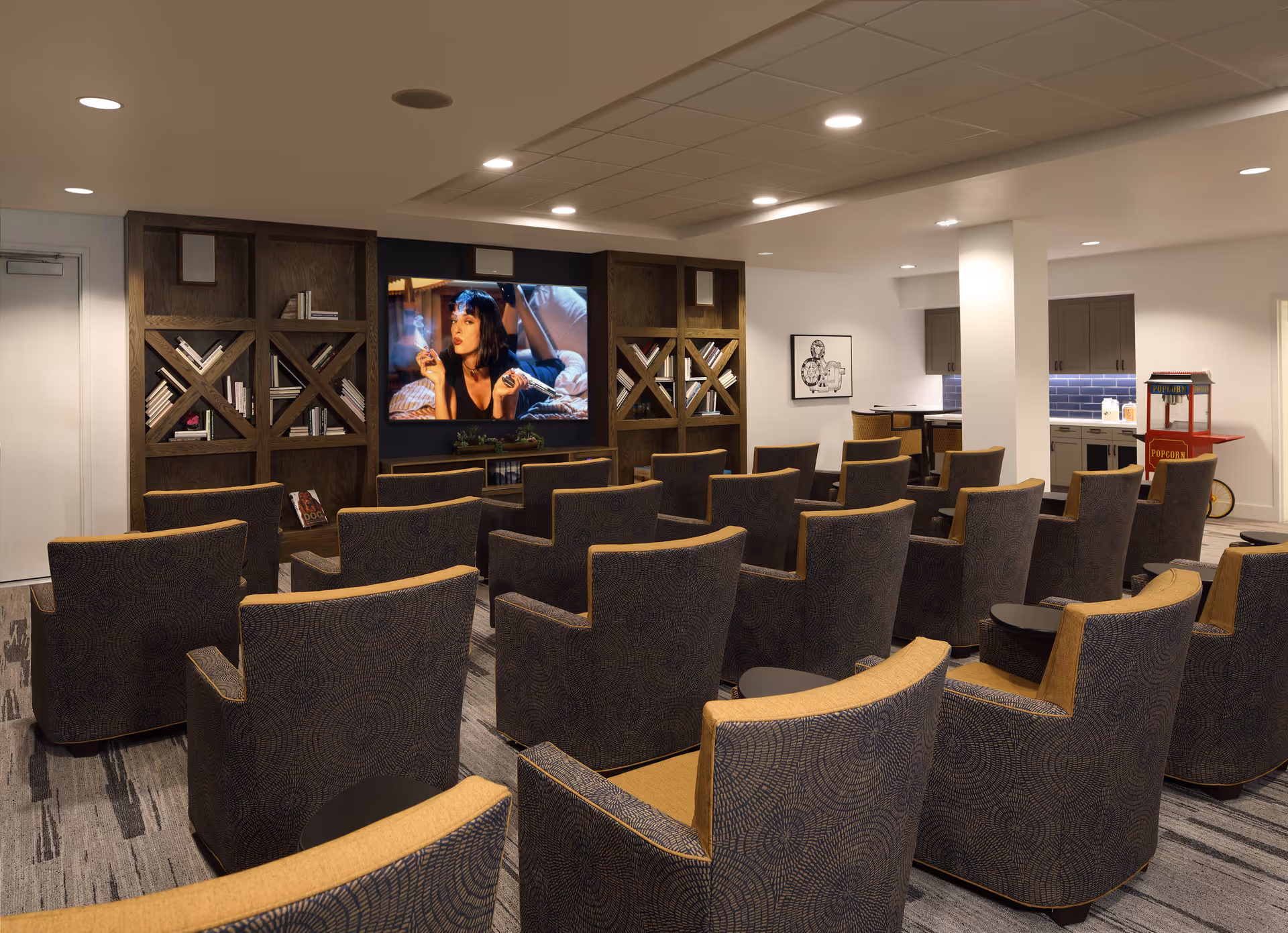A cozy senior living facility media room with multiple rows of patterned armchairs facing a large flat-screen TV mounted on a wooden bookshelf. The TV is showing a scene from a movie. To the right, there is a kitchenette area with cabinets, a countertop, and a popcorn machine. The room is well-lit with recessed ceiling lights.
