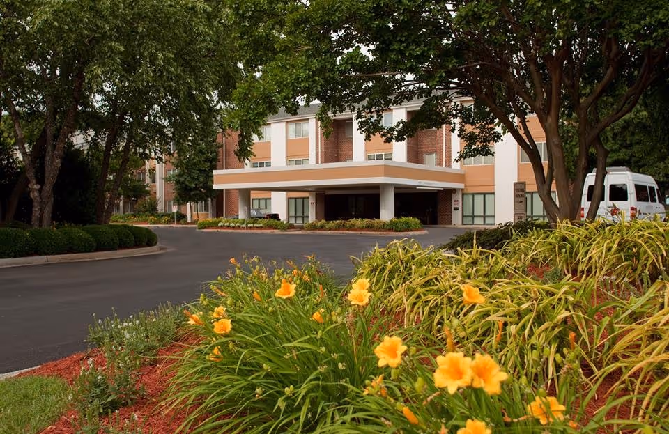 Entrance of a brick senior living building with a covered driveway and landscaped flower beds with yellow daylilies in the foreground.
