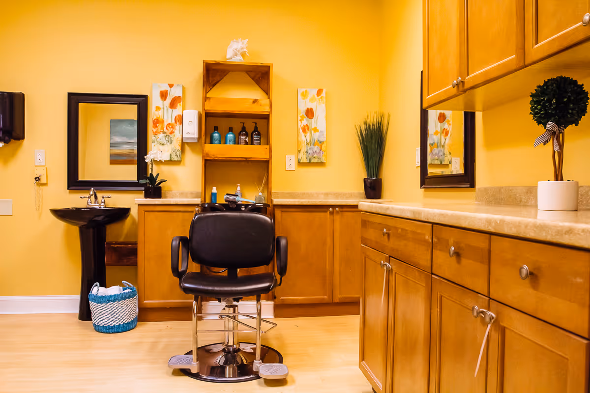 A salon-style hair station with a black barber chair, sink, mirrors, and wooden cabinets in a warm yellow room.