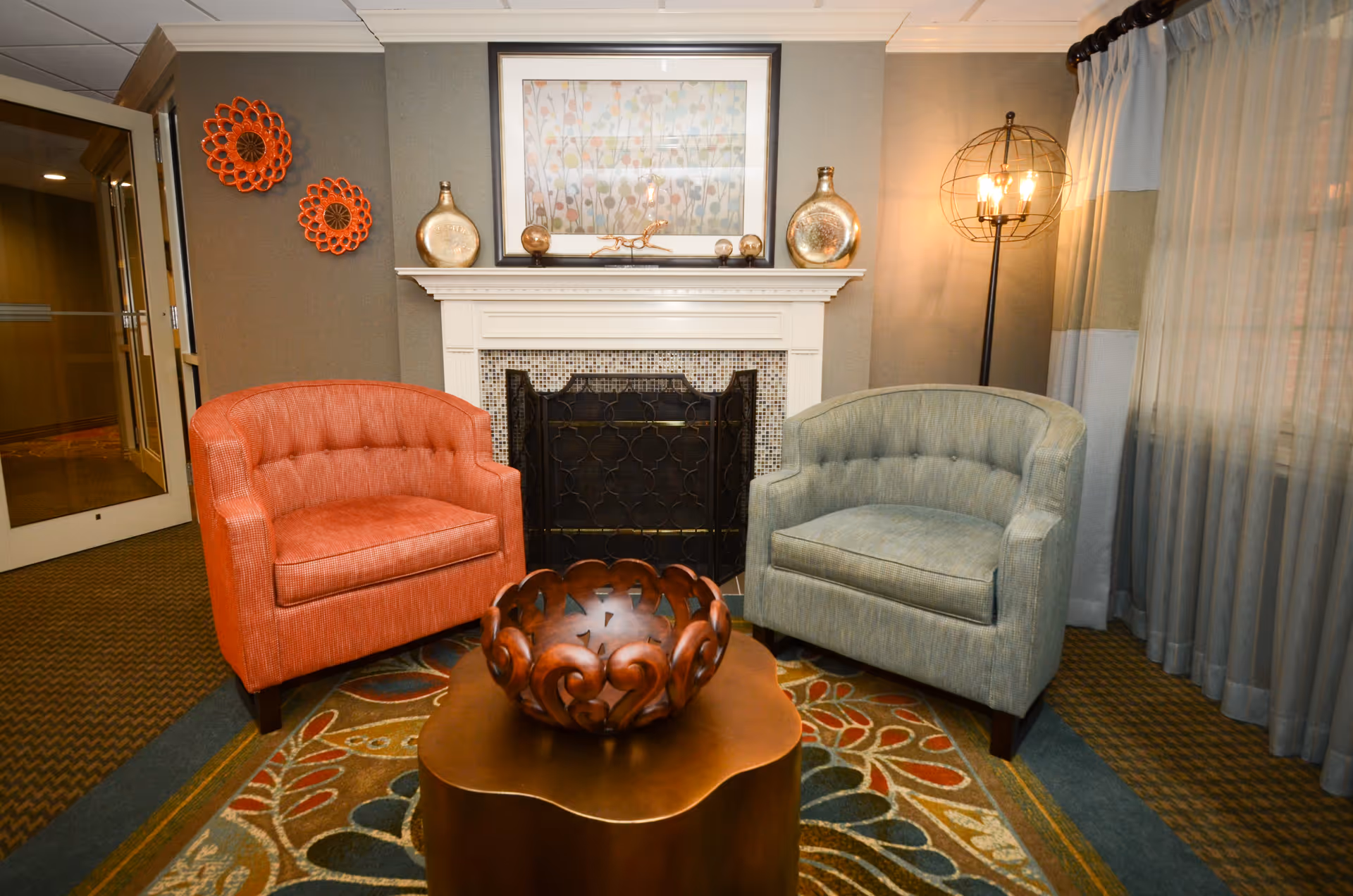 A cozy sitting area with a red armchair and a blue armchair placed on a patterned rug in front of a white fireplace. The fireplace mantel holds decorative vases and a framed floral artwork. To the right, there is a tall floor lamp with an exposed bulb design, and sheer curtains cover the window.