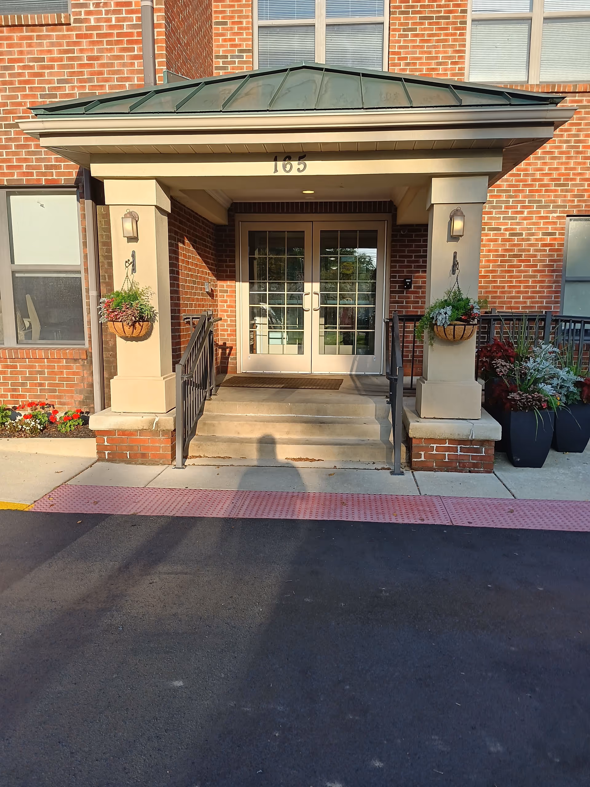 Brick building entrance with double glass doors, concrete steps, and hanging flower planters.