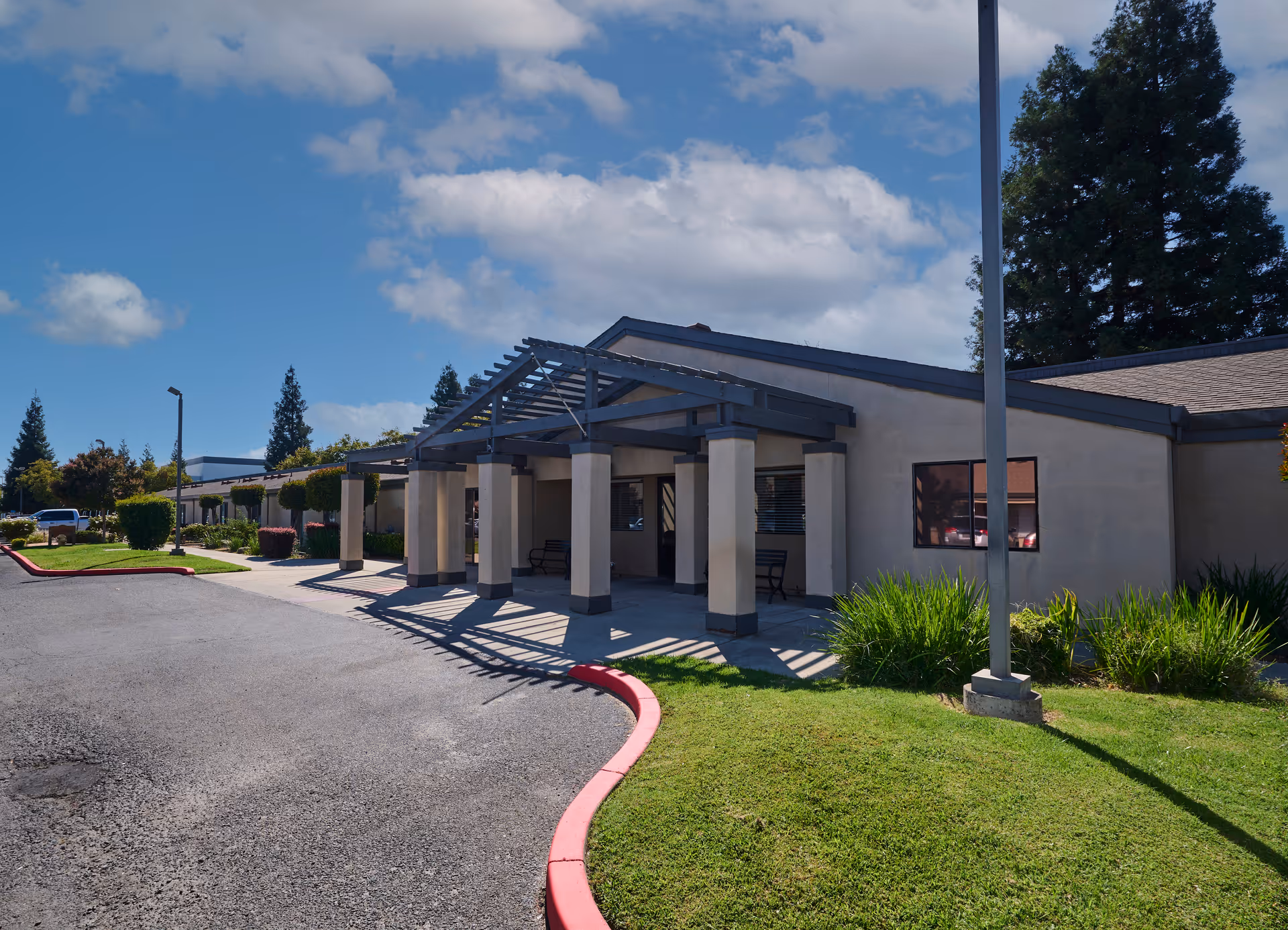 Front entrance of a single-story building with a covered pergola, columns, driveway, and landscaped lawn under a blue sky.