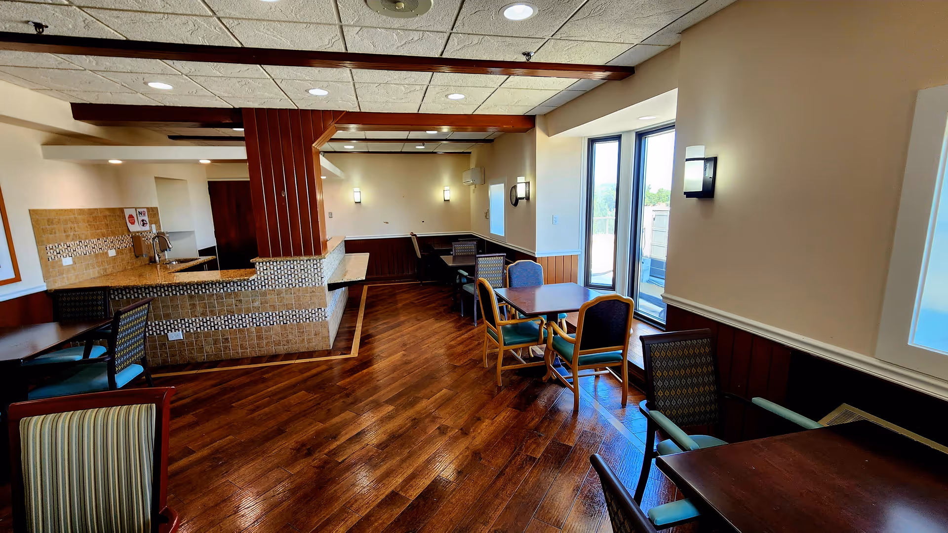 Interior common area with wooden floors, tables, chairs, and a counter in a senior living facility.