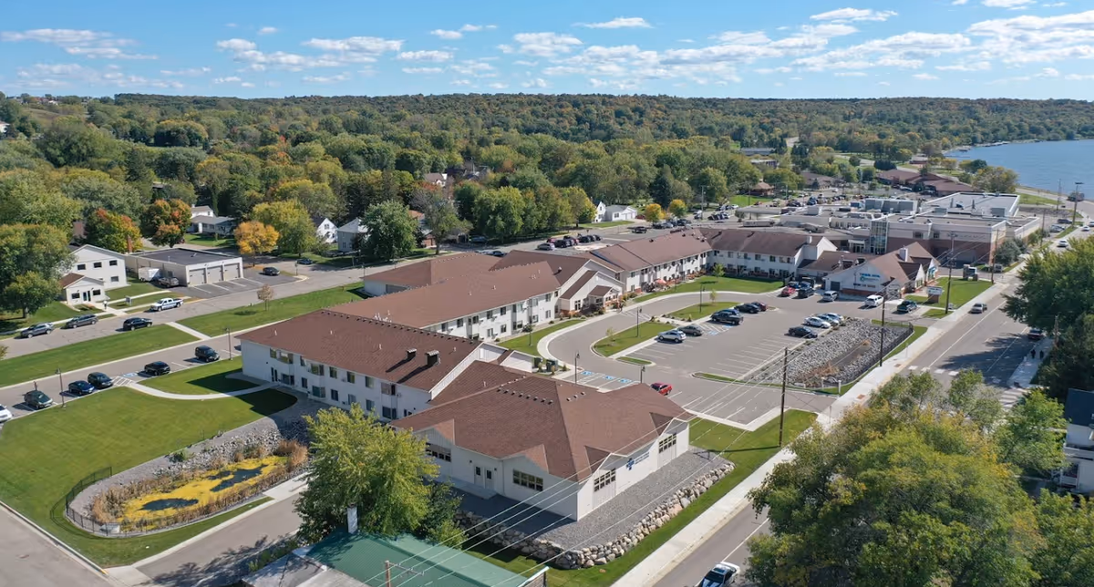 Aerial view of Ridgewood Villa Senior Living Apartments and surrounding area, showing multiple connected buildings with brown roofs, parking lots with cars, green lawns, trees, and a body of water in the background under a partly cloudy sky.