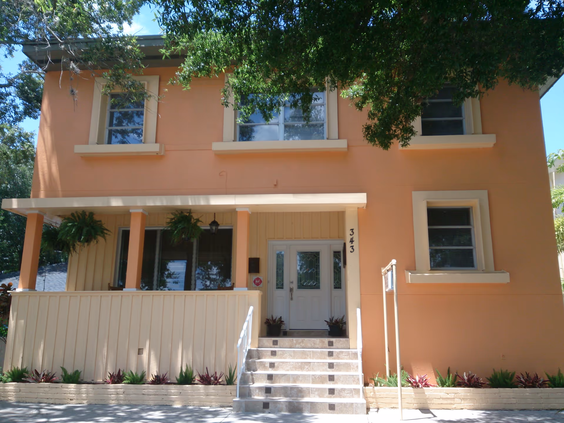 Front view of a two-story peach-colored building with a small porch, steps leading to double doors, and hanging plants.