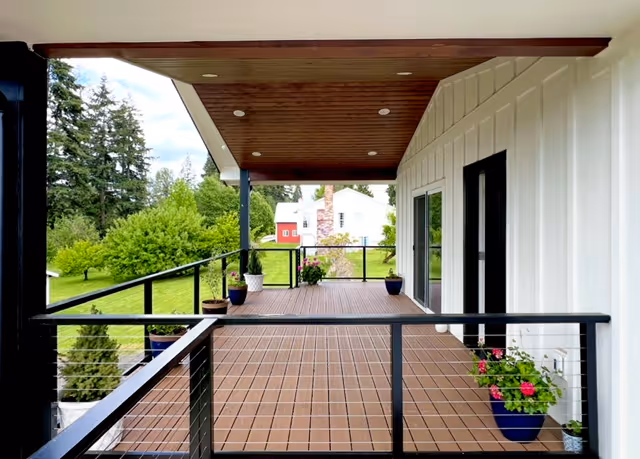 Outdoor wooden balcony with planters extends from white building, railing and lawn beyond.