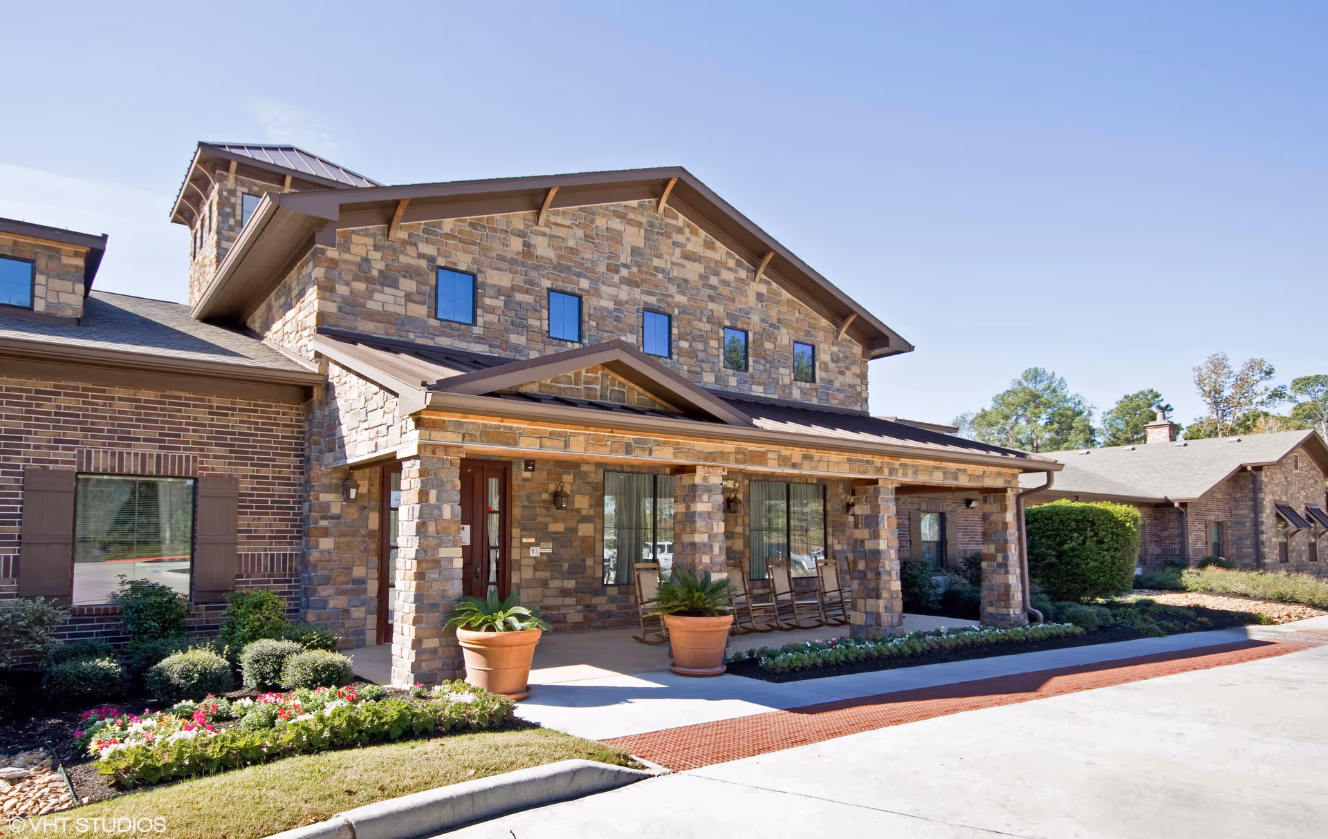 Exterior view of a two-story assisted living facility building with stone and brick facade, a covered porch with rocking chairs, potted plants, and landscaped flower beds under a clear blue sky.