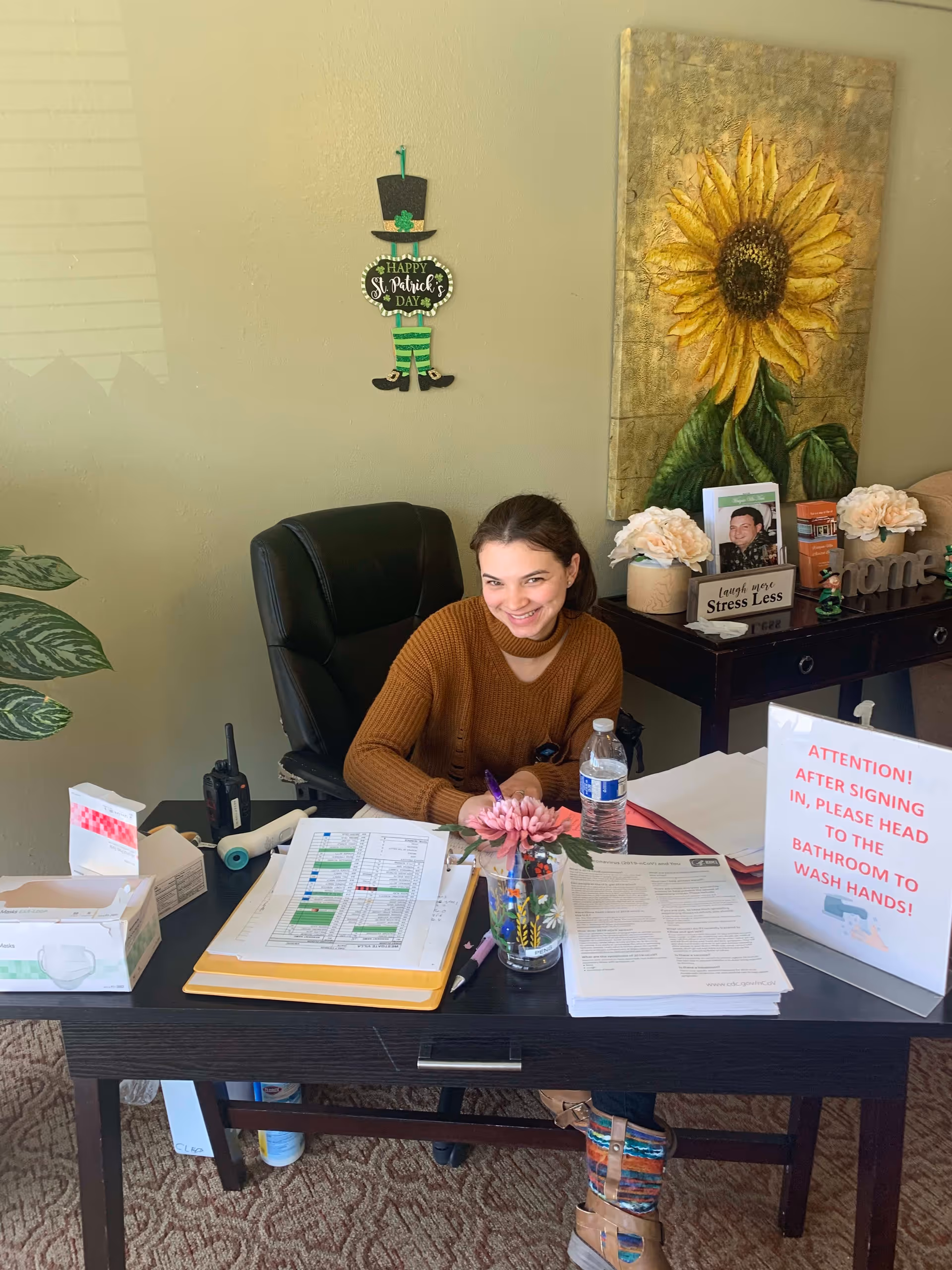 A woman sitting at a desk in an office area, smiling and writing on a clipboard. The desk has various papers, a water bottle, a walkie-talkie, and a sign that reads 'ATTENTION! AFTER SIGNING IN, PLEASE HEAD TO THE BATHROOM TO WASH HANDS!'. Behind her is a wall with a sunflower painting, a St. Patrick's Day decoration, and a table with flowers and framed photos.