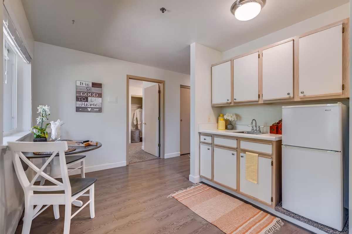 Compact kitchen with white cabinets, sink, fridge, and a small dining table by a window.