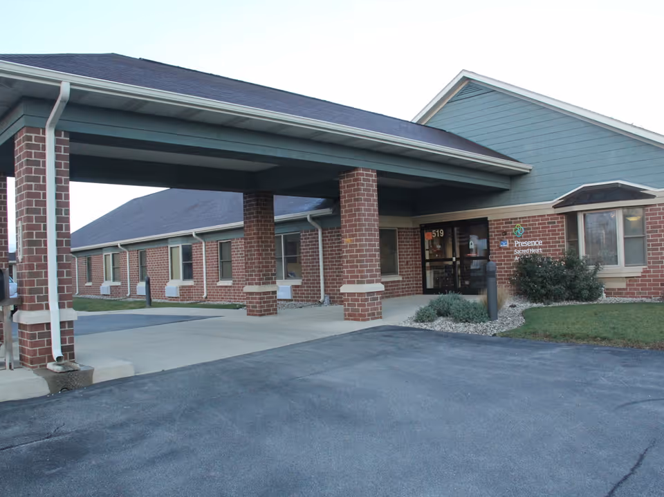 Covered entrance and driveway of a single-story brick assisted living building with a 'Presence Sacred Heart' sign.