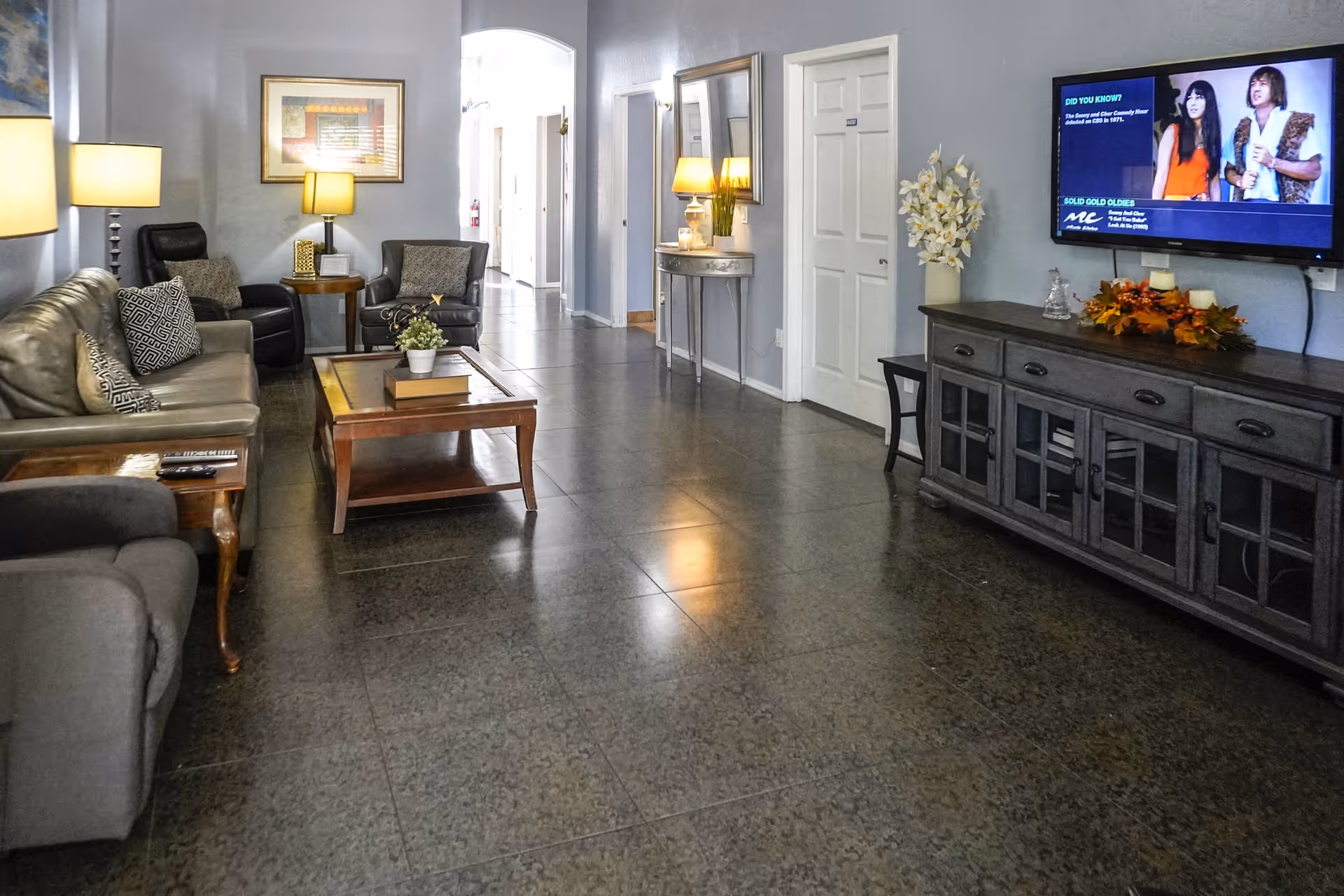 Spacious senior living lounge with seating, coffee table, TV, and hallway leading to rooms.