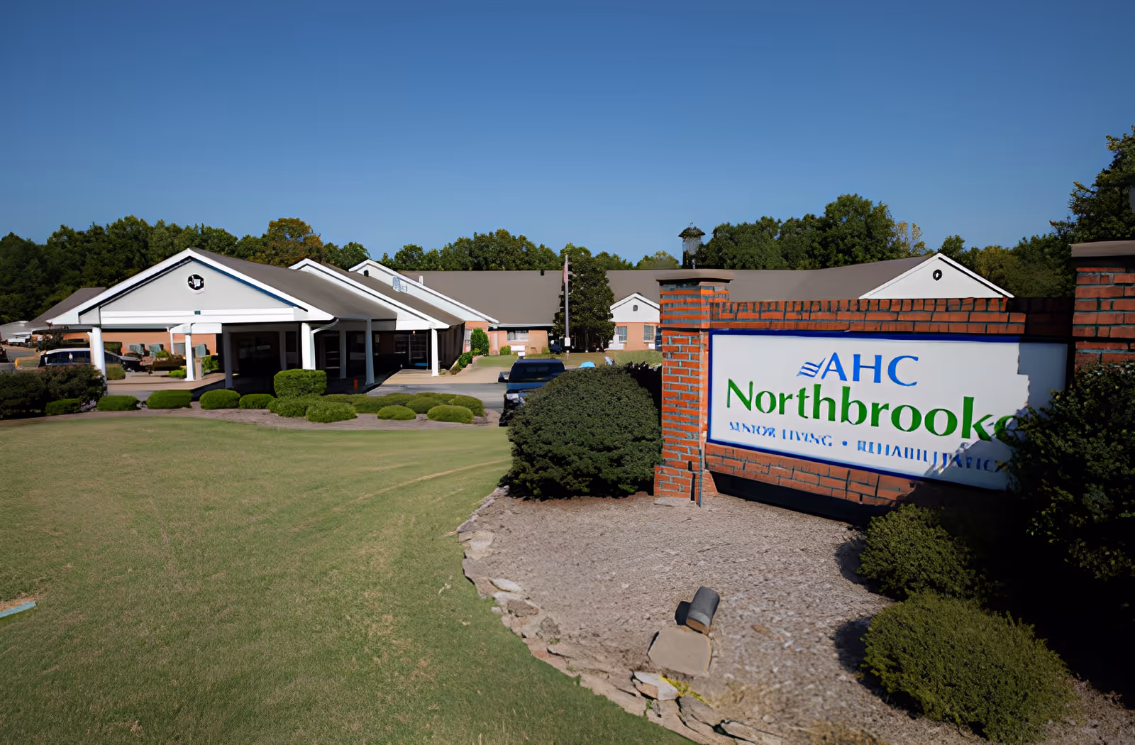 Exterior view of a senior living facility with a large green lawn and neatly trimmed bushes. The building has a white entrance canopy with columns and a beige roof. A brick sign in the foreground reads 'AHC Northbrooke Senior Living - Rehabilitation'. Trees are visible in the background under a clear blue sky.