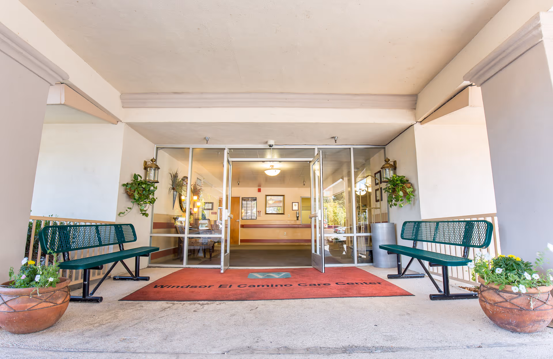 Entrance area of Windsor El Camino Care Center showing a covered walkway with two green metal benches on either side, potted plants, and glass double doors leading into the facility's lobby.