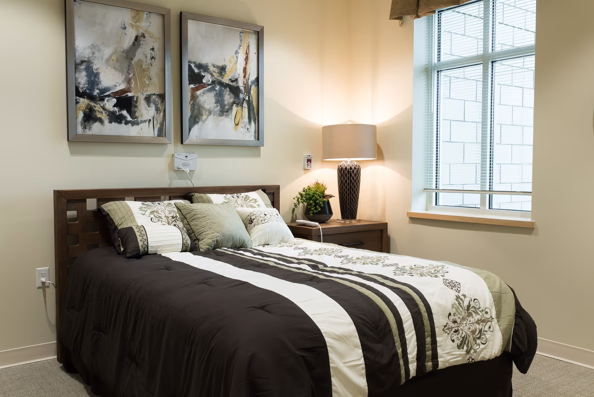 A neatly made bed with a dark brown, white, and green patterned comforter and multiple pillows in a well-lit bedroom. There are two abstract paintings hanging on the wall above the headboard. A wooden nightstand with a decorative lamp and a small plant is positioned next to the bed. A window with blinds is on the right side of the room.