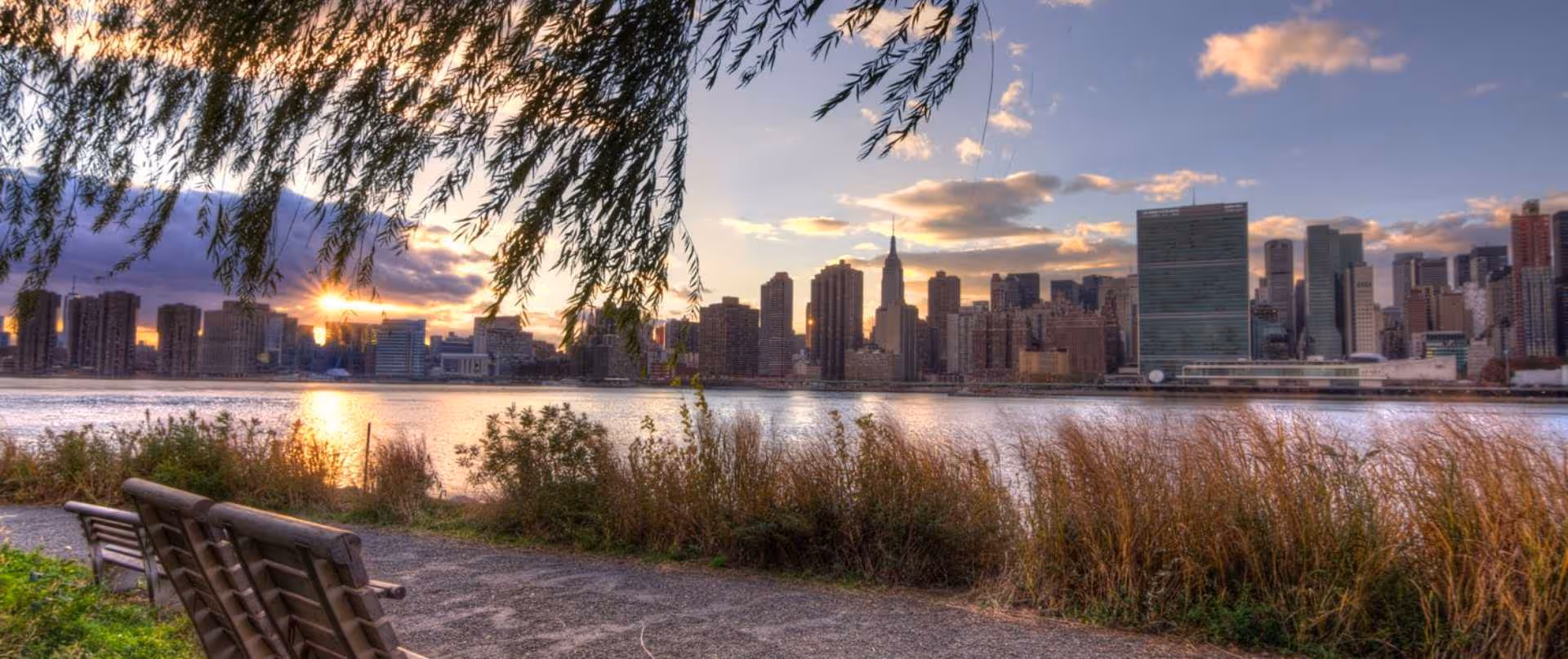 Riverside park bench overlooks a city skyline at sunset.