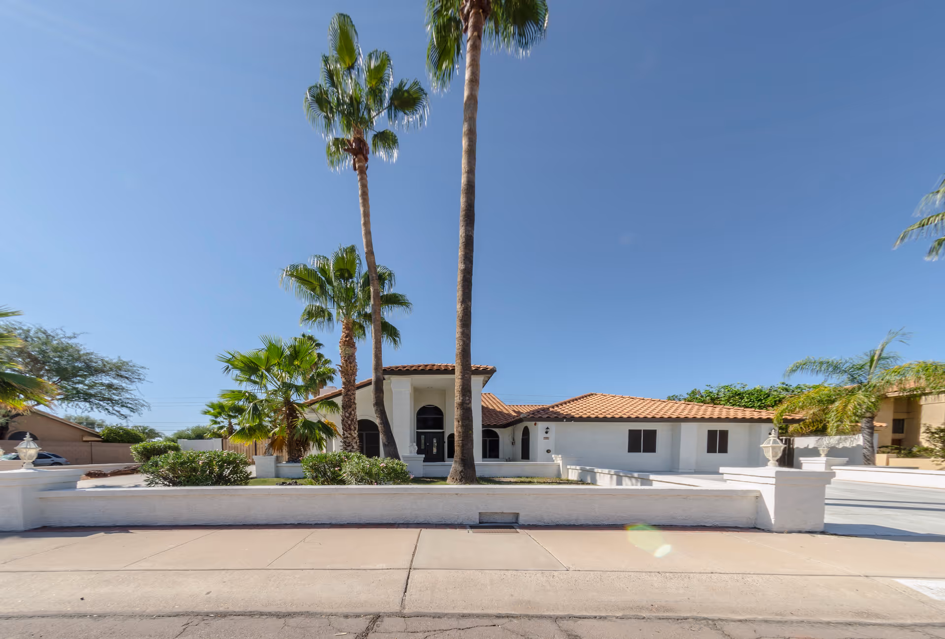 Front exterior view of a single-story building with white walls and a red-tiled roof, surrounded by palm trees and a low white wall, under a clear blue sky.