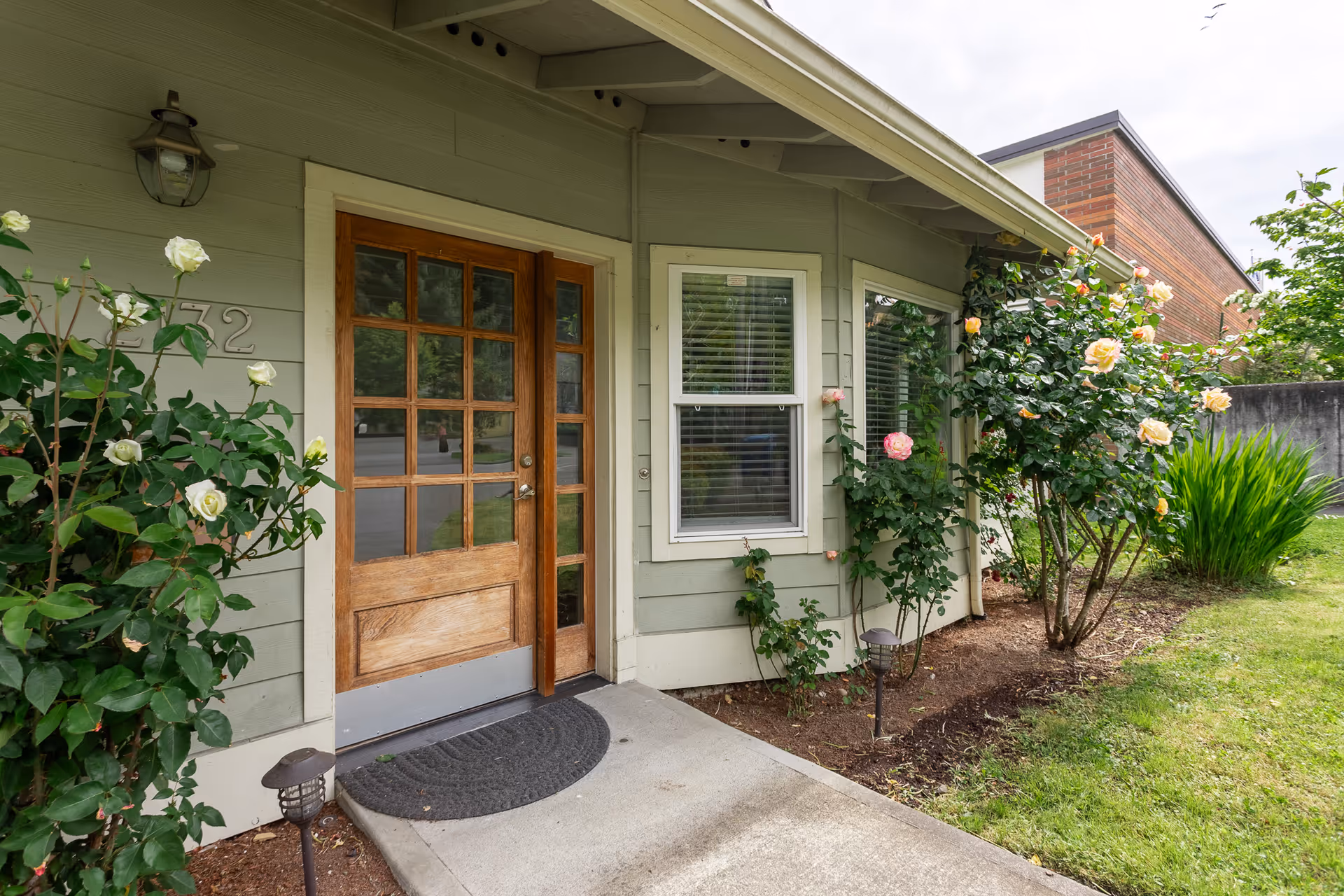 Green exterior home with wooden front door, windows, and blooming roses along the walkway.