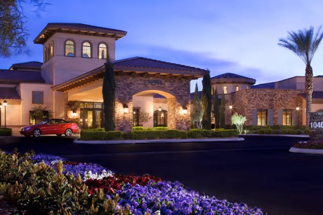 Dusk view of a Mediterranean-style building entrance with an arched porte-cochère, illuminated windows, landscaped flower beds, and a parked red car.