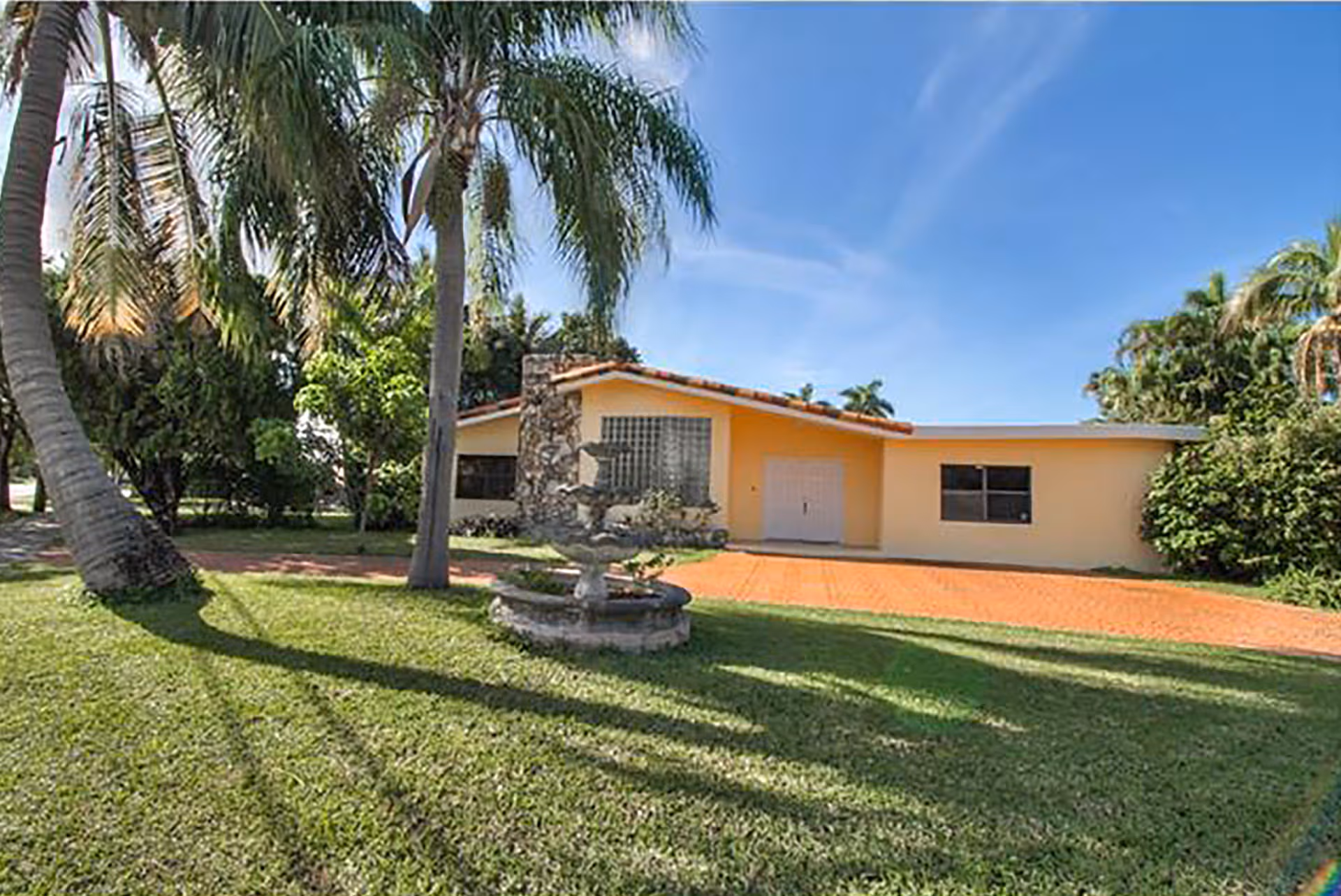 Sunlit peach-colored, single-story building with palm trees and a circular fountain in front.