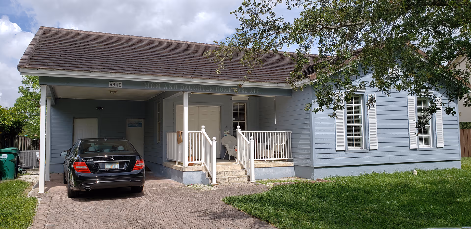 Front exterior view of a single-story light blue house with white trim and shutters. There is a small porch with white railings and two chairs. A black car is parked in the driveway under a covered carport. The house number 9640 is visible above the porch. The sky is partly cloudy and there is green grass and a tree in the yard.