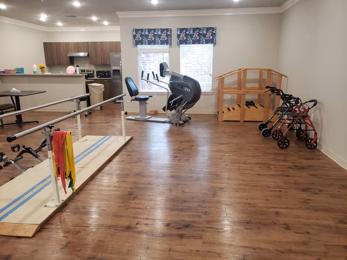 Indoor rehabilitation gym with parallel bars, an exercise bike, and walkers near a kitchenette.