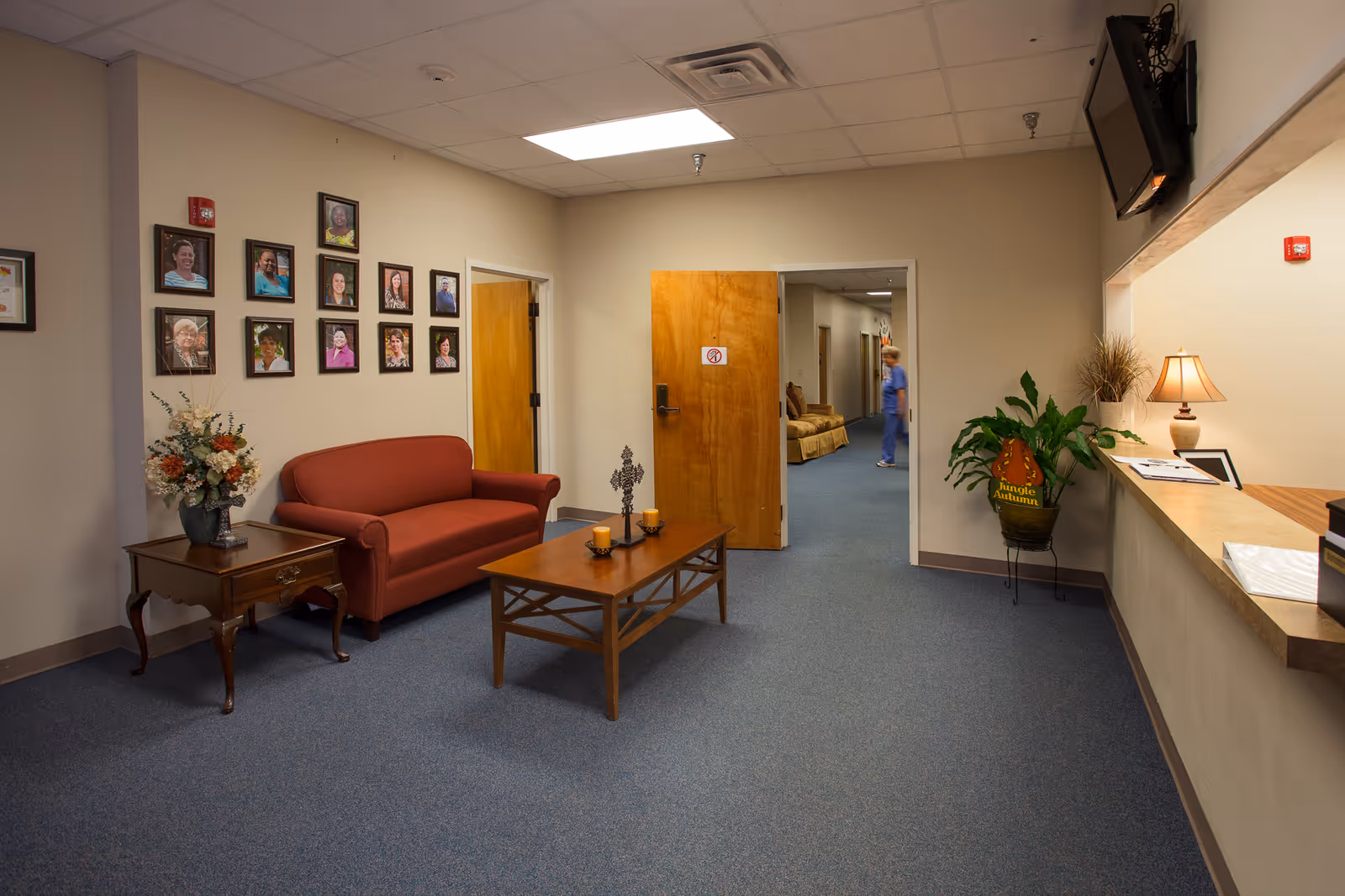Lobby seating area with a red sofa, coffee table, wall portraits, plants, and a reception counter.