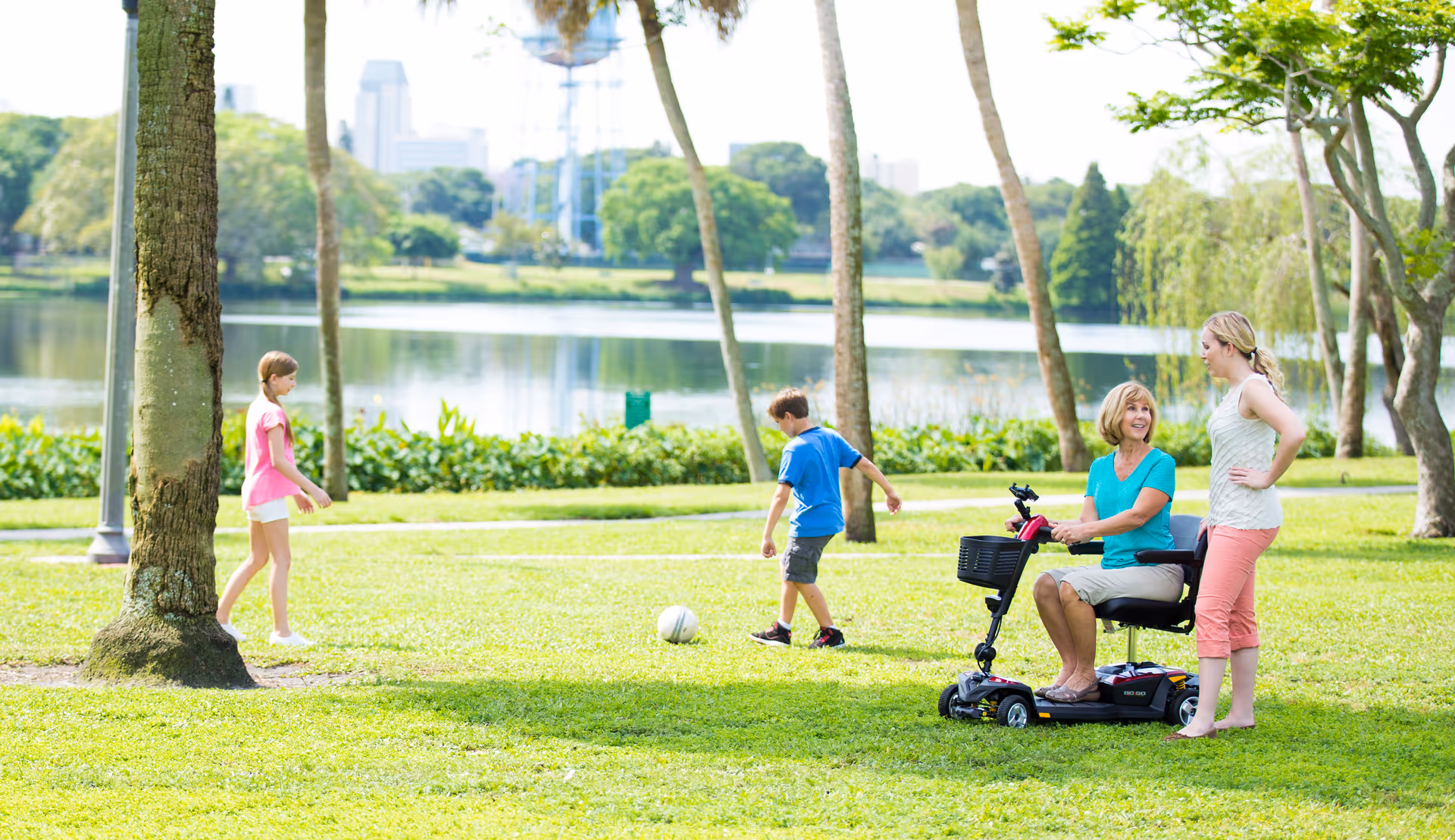 Senior on mobility scooter outdoors as caregiver and children play in a park.