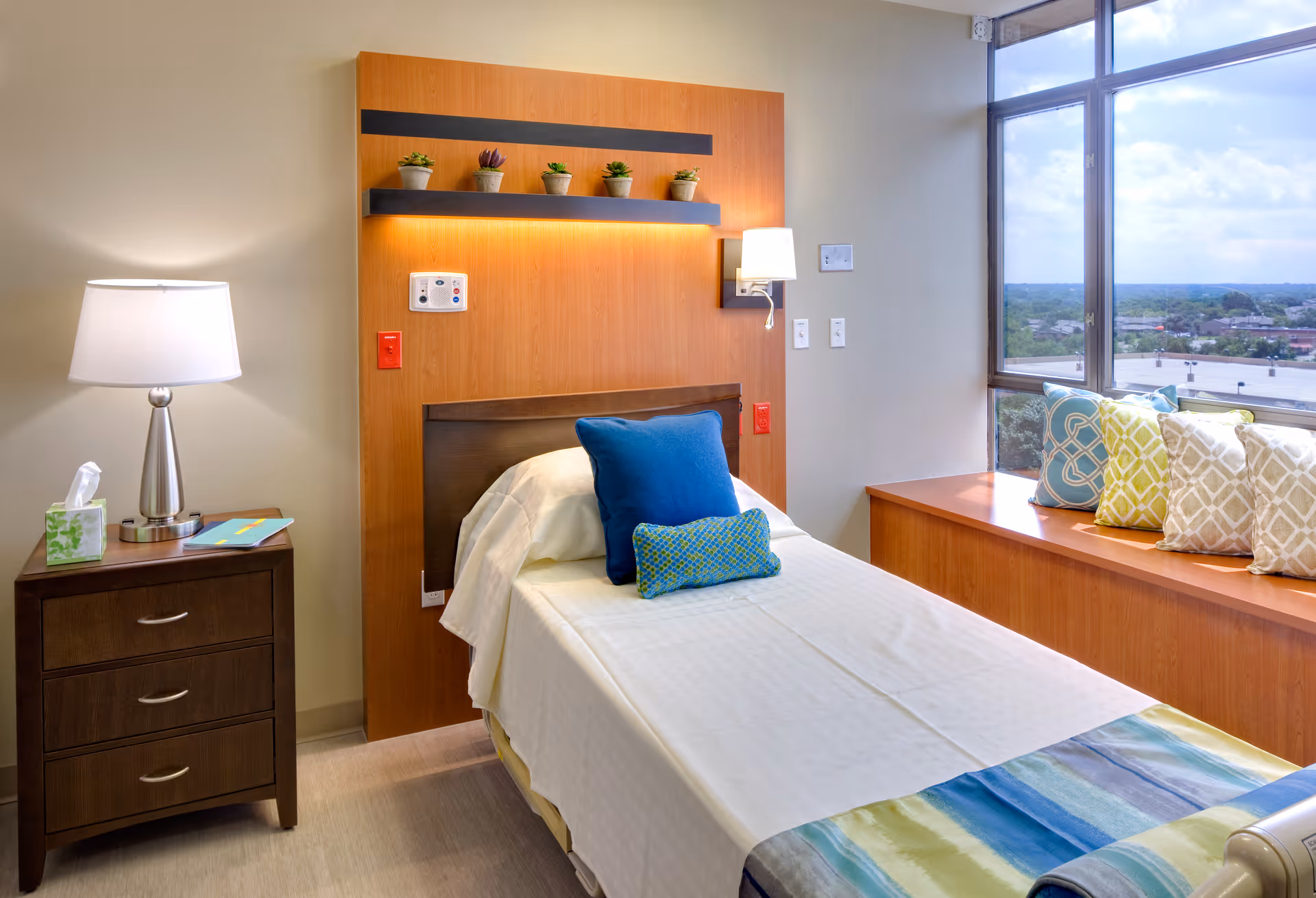 A modern hospital-style bedroom with a bed, nightstand, lamps, and a windowed seating area.