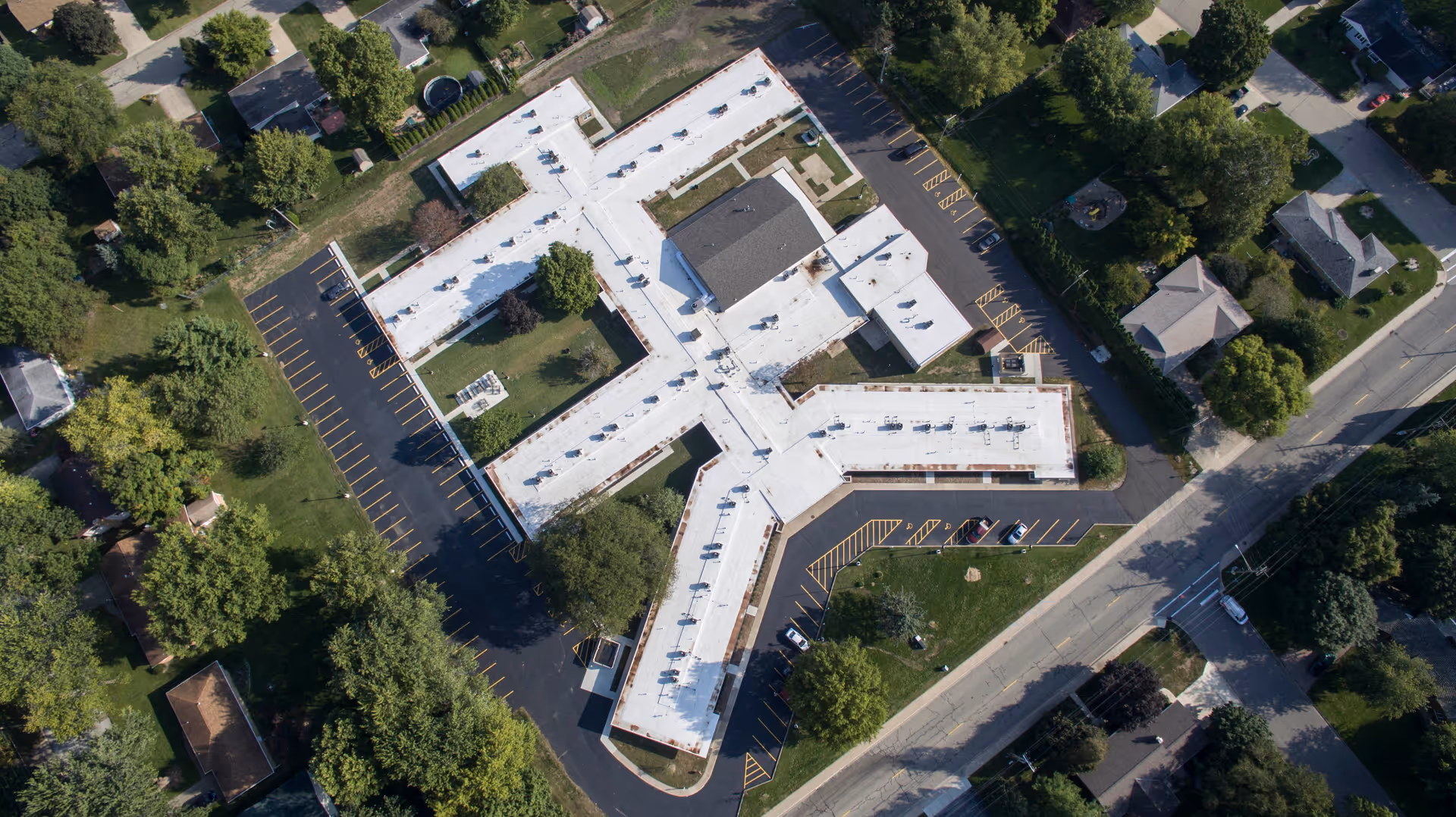 Aerial view of Morton Estates senior living facility showing a large, white-roofed building with multiple wings surrounded by parking lots, trees, and nearby residential houses.