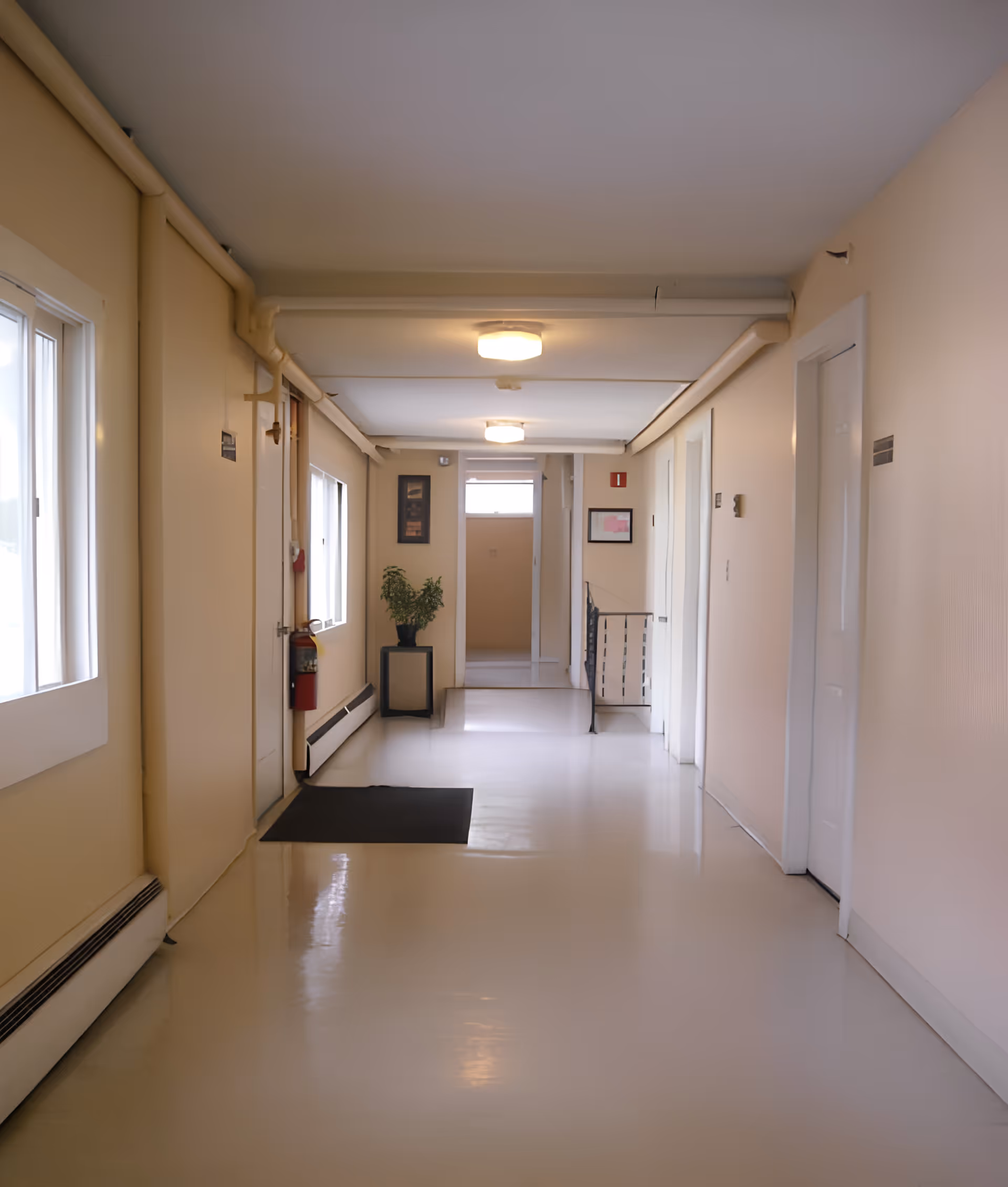 A clean, well-lit hallway in a senior living facility with beige walls, white doors, and a shiny floor. There are two ceiling lights, a fire extinguisher mounted on the left wall, a small black mat on the floor, a potted plant on a black stand near the end of the hallway, and framed pictures on the walls.