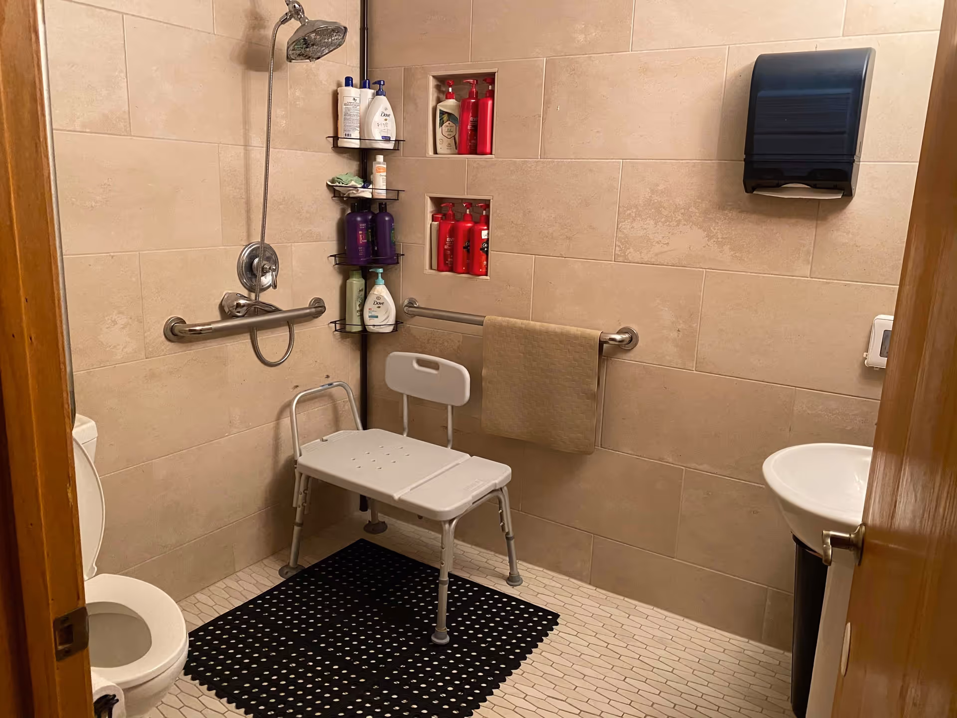 Accessible bathroom with shower chair, grab bars, toilet, sink, and supplies.