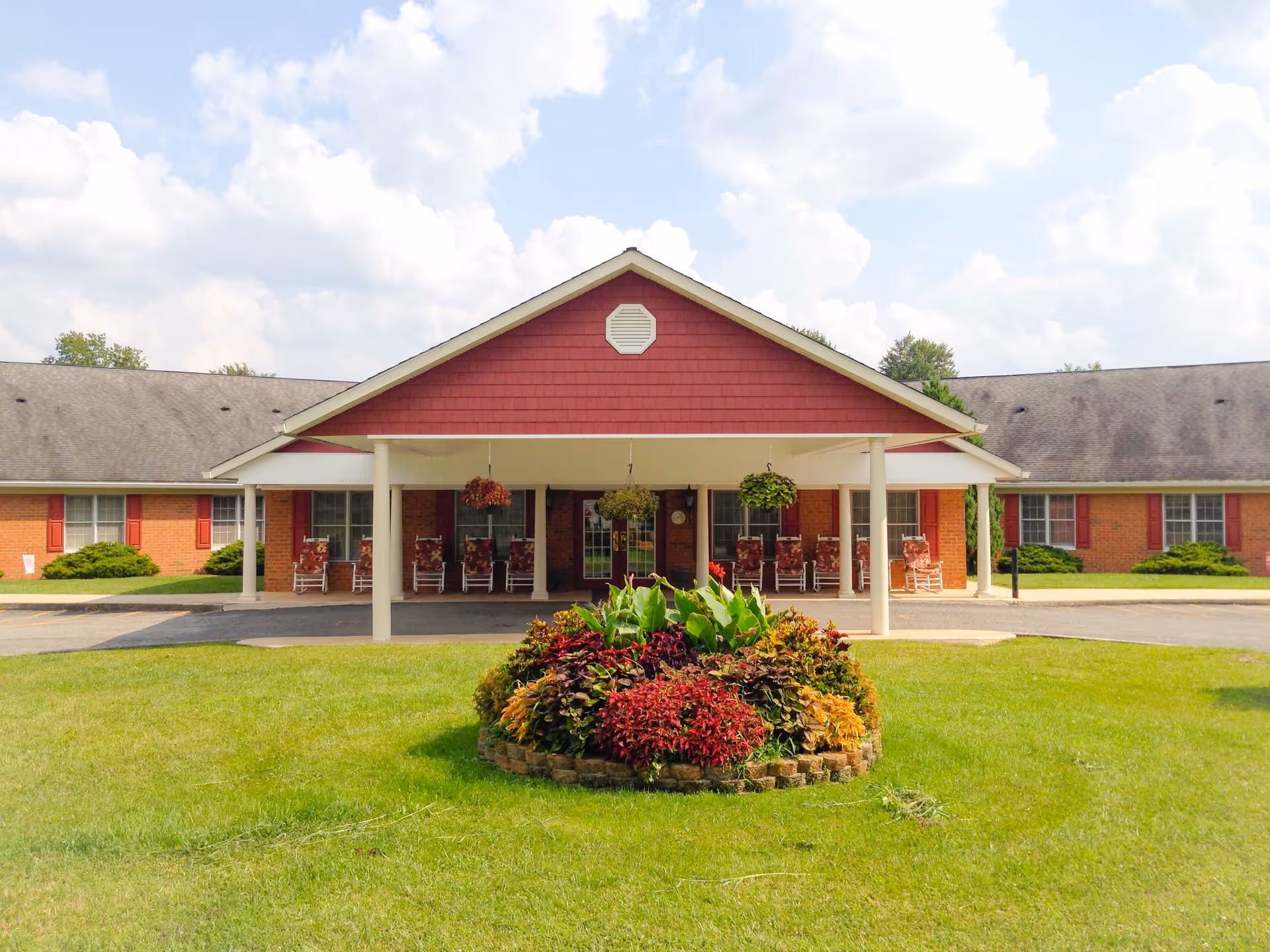 Front exterior view of a single-story assisted living and retirement center building with a covered entrance supported by white columns. The building has red siding and brick walls with multiple windows and red shutters. There is a circular flower bed with colorful plants and greenery in the foreground, and several rocking chairs are placed under the covered entrance.