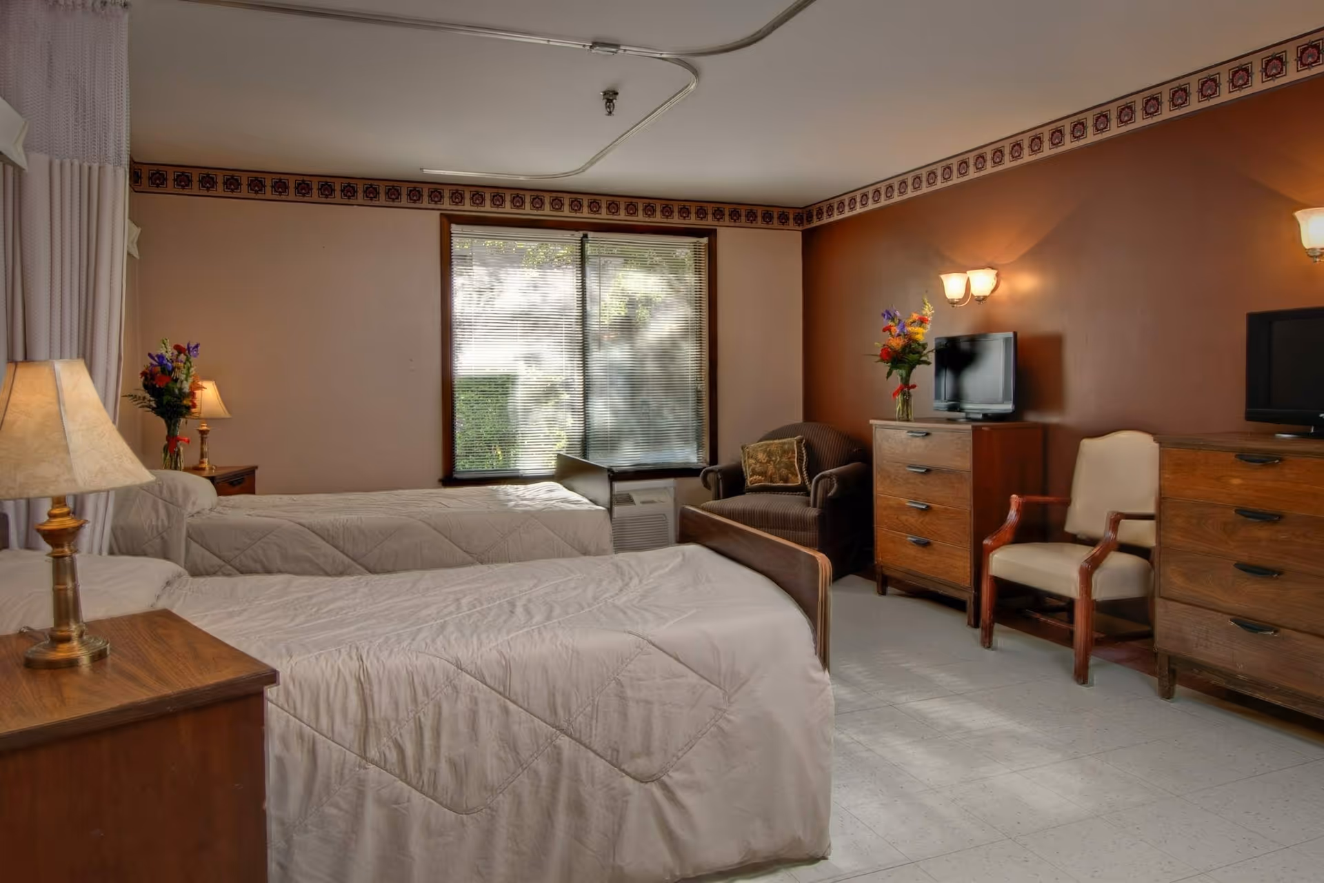 A nursing facility bedroom with two single beds covered in beige quilts, a wooden nightstand with a lamp and a vase of flowers, a window with blinds, a brown armchair with a decorative pillow, two wooden dressers with small flat-screen TVs on top, and a wooden chair with armrests. The walls are painted beige and brown with a decorative border near the ceiling.