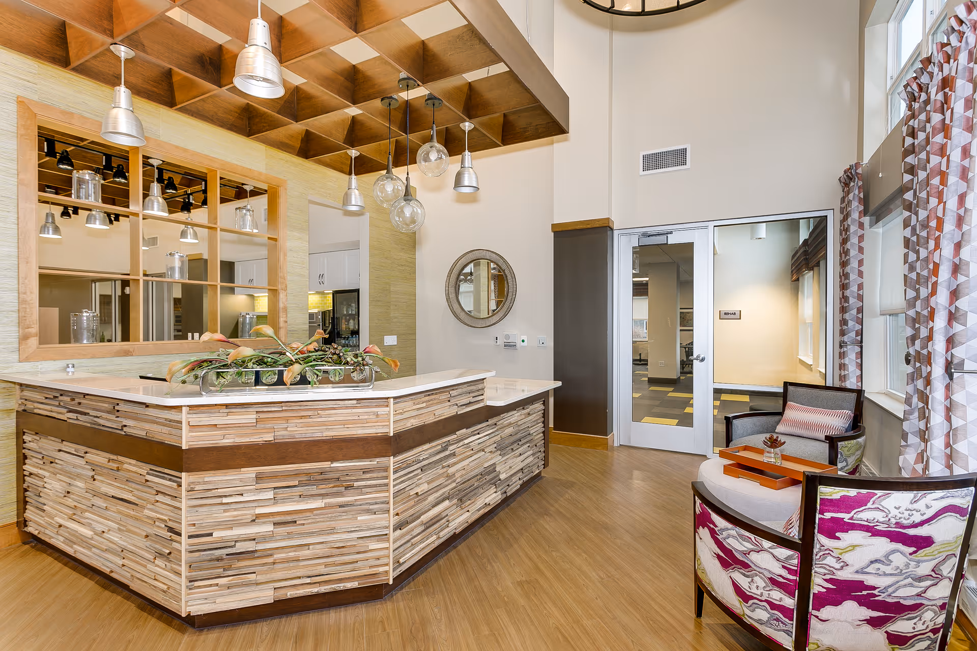 A modern reception area with a wooden textured front desk topped with a white countertop and a decorative plant. Behind the desk is a large window with wooden framing and hanging pendant lights. To the right, there is a small seating area with two armchairs, a round ottoman, and a small table with a decorative item. The room has large windows with patterned curtains, light-colored walls, and a wooden floor.