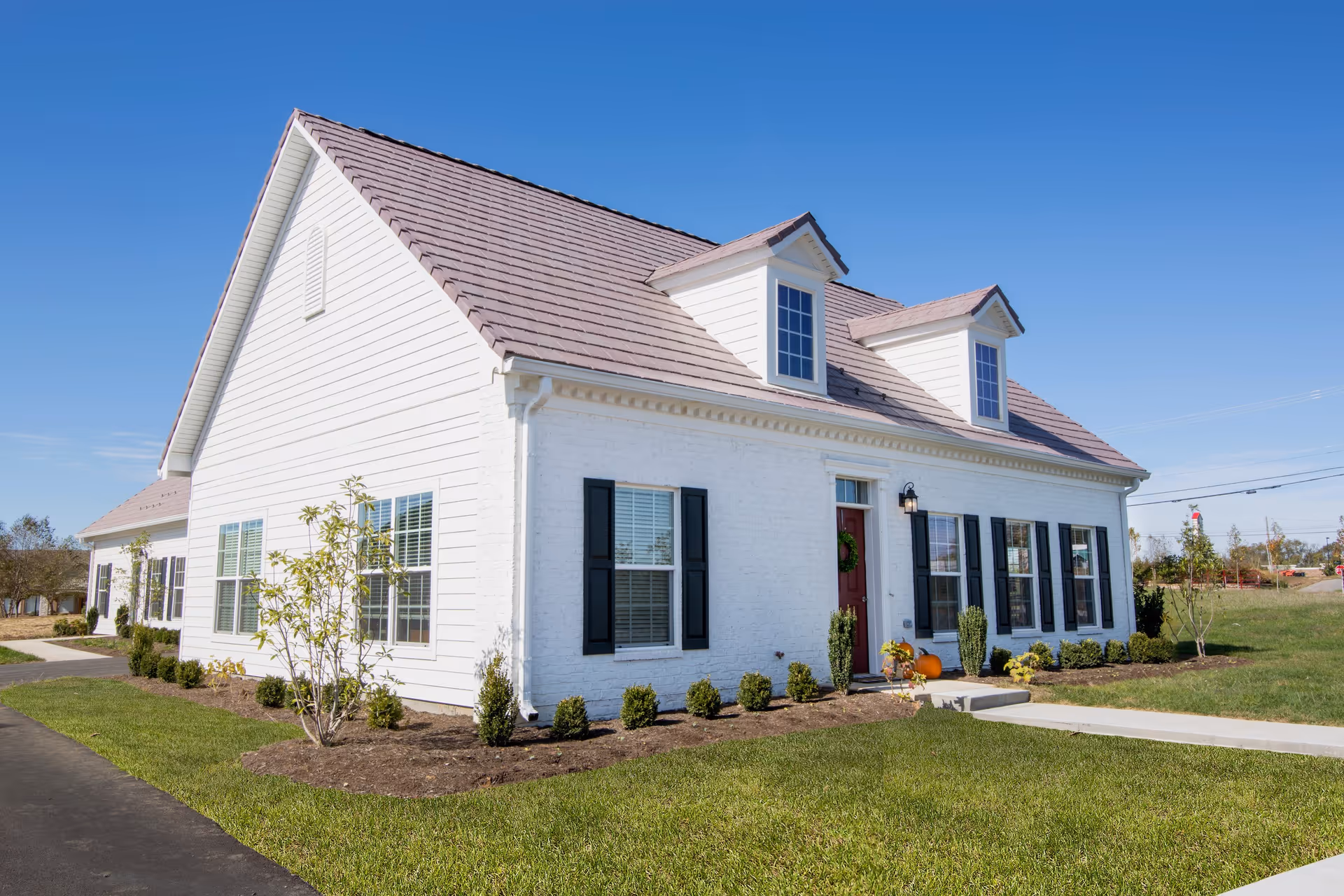 White brick building with dark shutters, a red roof, and dormer windows on a sunny day.
