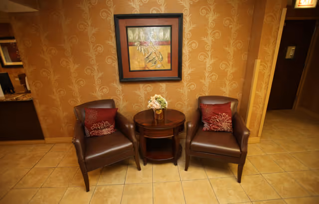 Two brown leather armchairs with red floral-patterned pillows are positioned on either side of a small round wooden table with a floral arrangement in a vase. The setting is against a wall with gold patterned wallpaper and a framed abstract painting above the table. The floor is tiled in a light beige color.