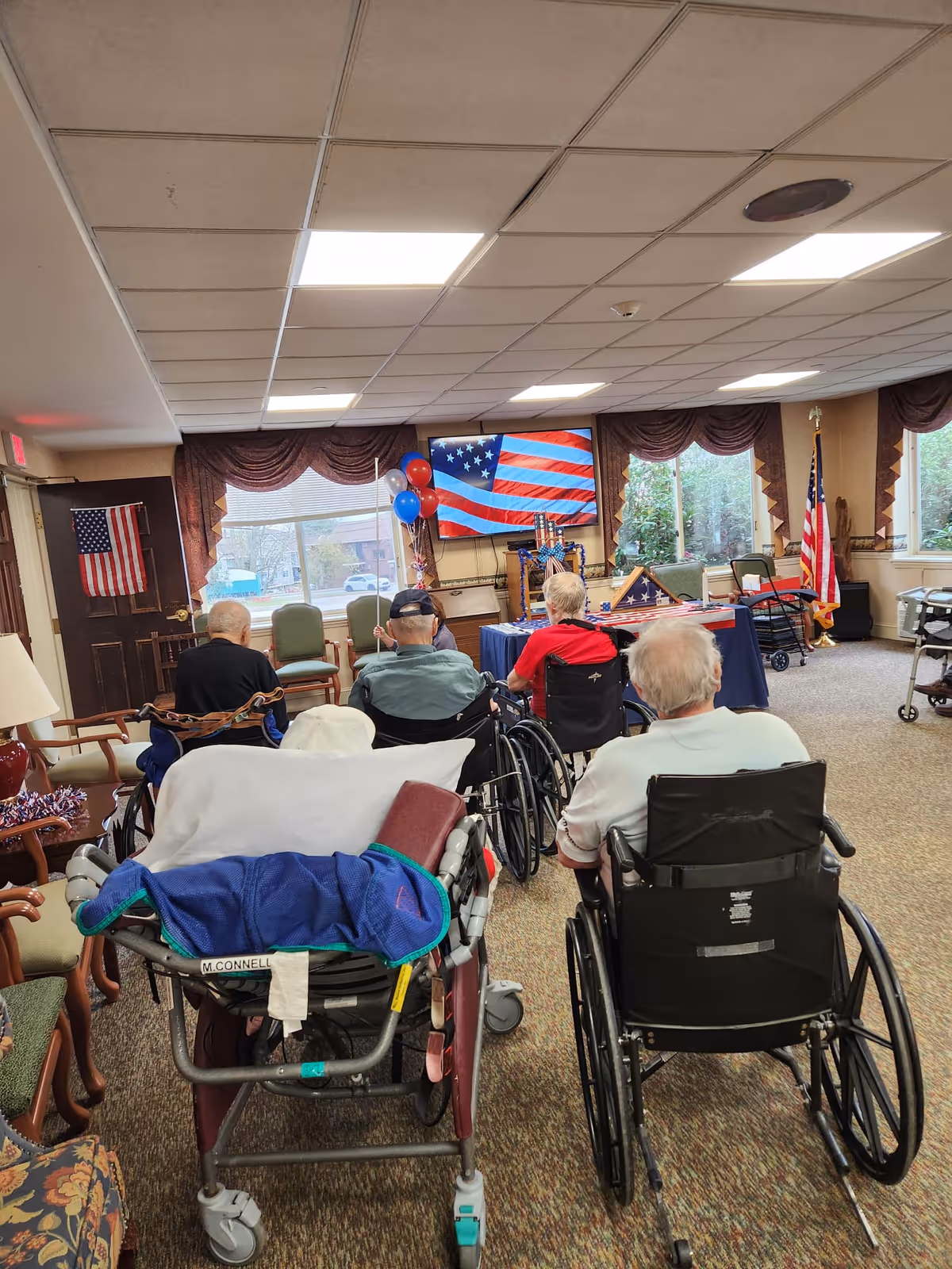 A group of elderly people in wheelchairs facing a television screen displaying an American flag in a decorated common room. The room has large windows with curtains, American flags, and patriotic decorations including balloons and folded flags on a table.