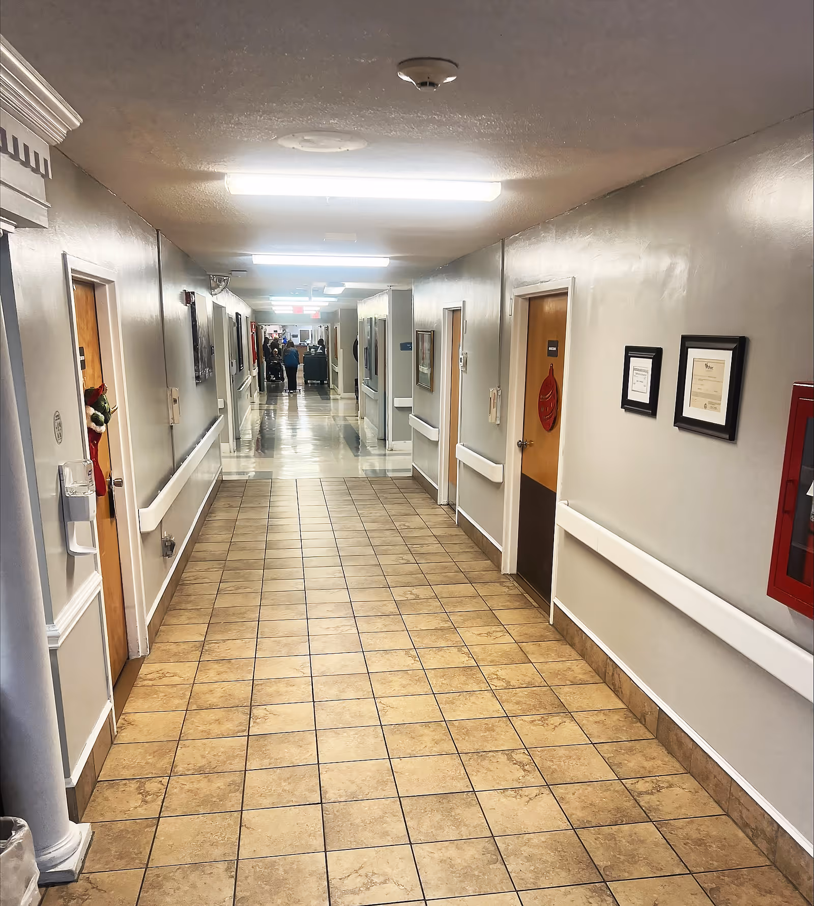 A long, clean hallway in a healthcare facility with tiled flooring and white walls. Several wooden doors line both sides of the corridor, some decorated with holiday items. Handrails run along the walls, and framed certificates or pictures are hung on the right side. Fluorescent lights illuminate the hallway, and a few people are visible in the distance near the end of the corridor.